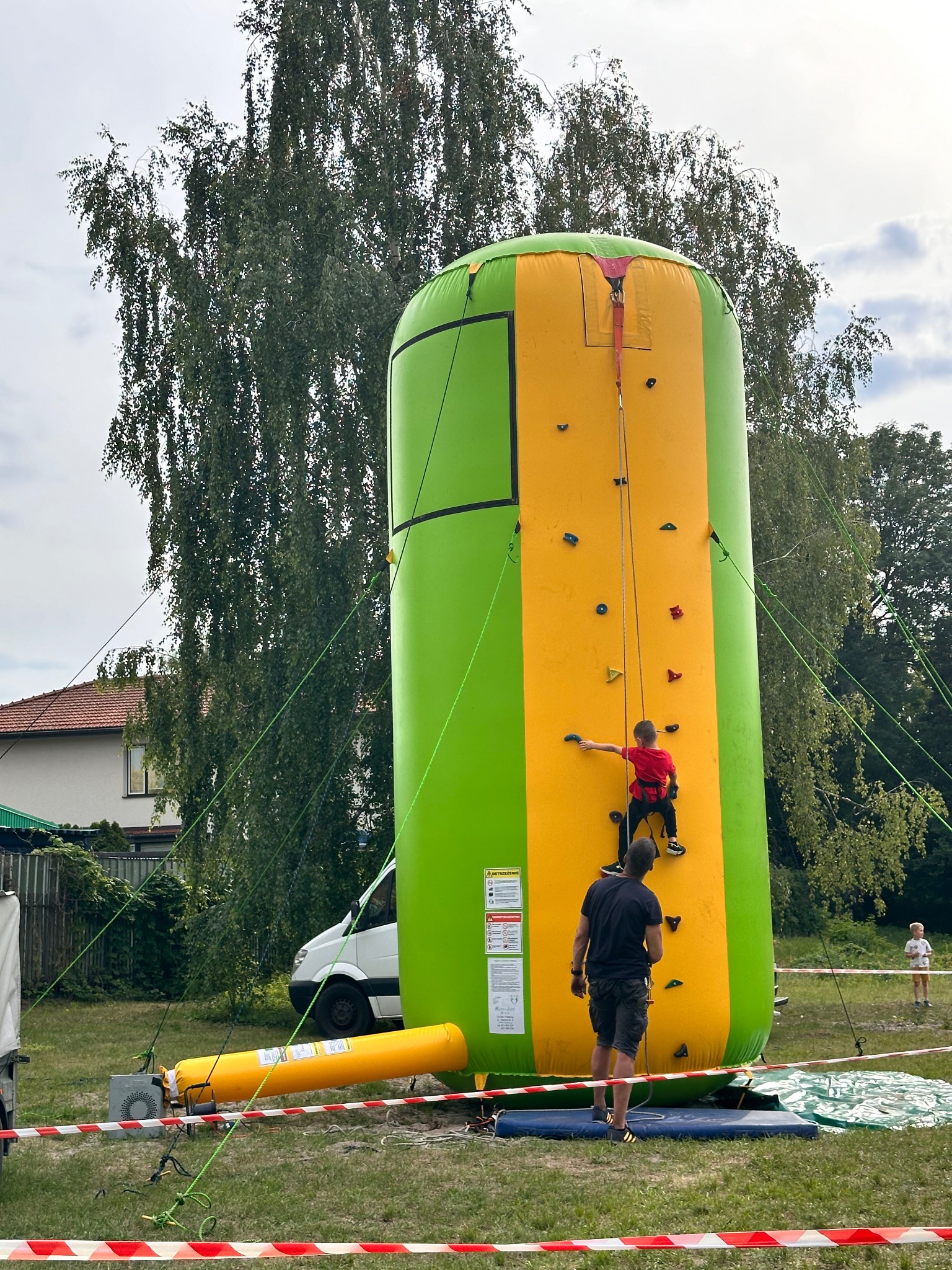 Dmuchana ścianka wspinaczkowa w kolorach zieleni i żółci, z chłopcem wspinającym się pod nadzorem instruktora. W tle drzewa i budynek.