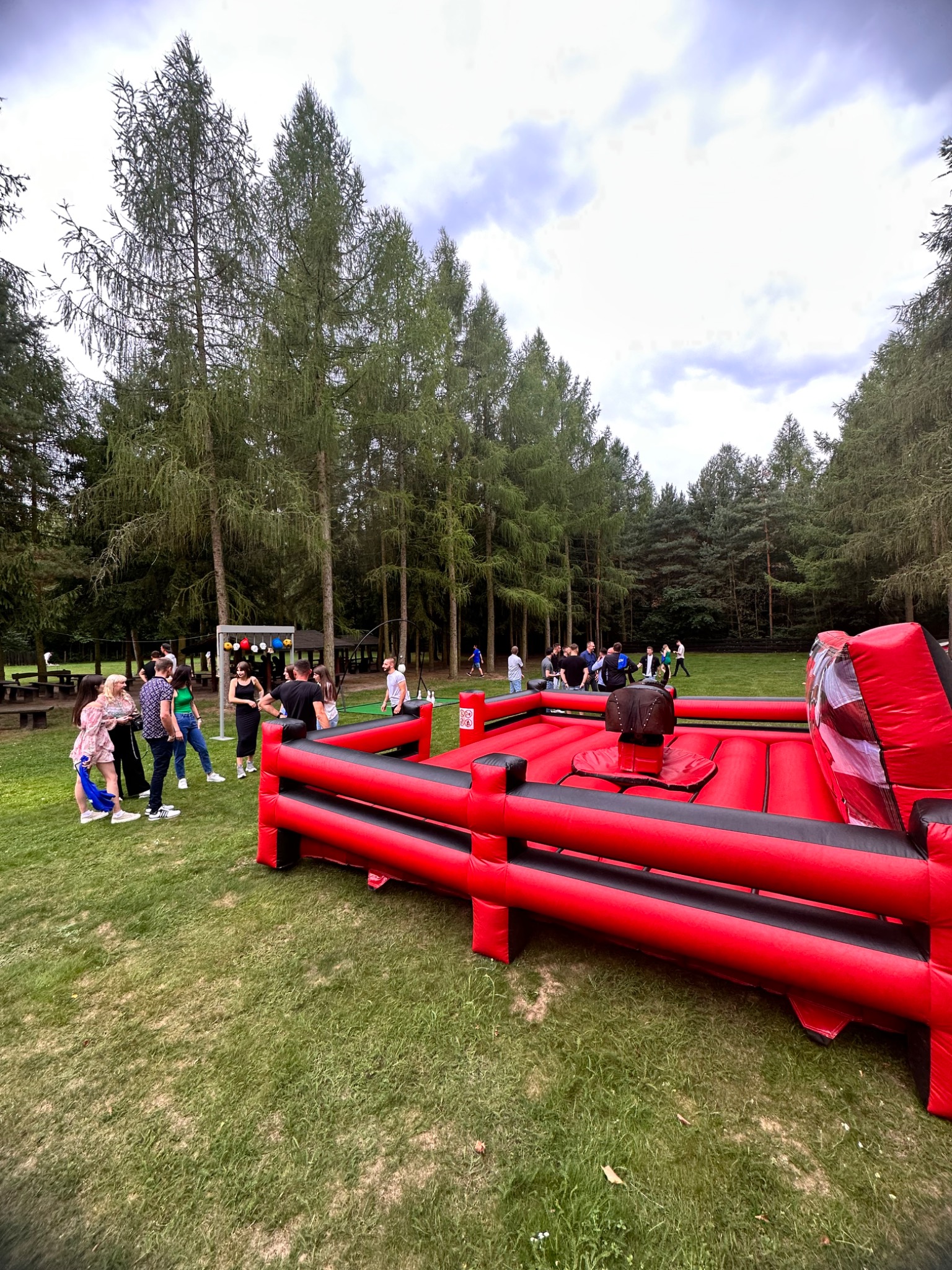 Czerwono-czarny dmuchany byk na zielonej trawie, w tle grupa ludzi i wysokie drzewa, elementy dekoracyjne