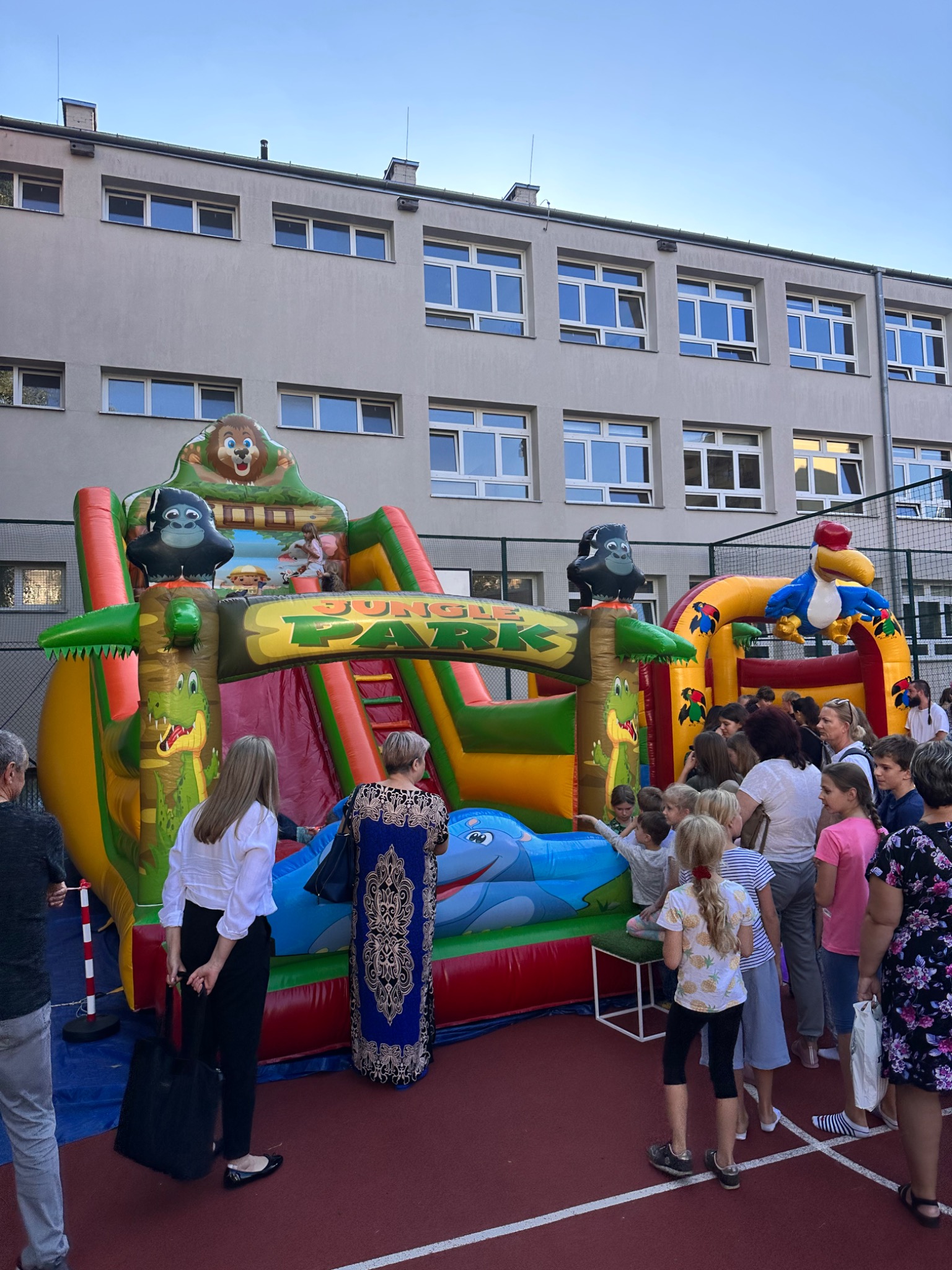 Kolorowy dmuchany plac zabaw z motywami dżungli, w tym krokodyle, goryle i papuga, otoczony grupą dzieci i dorosłych na tle budynku.