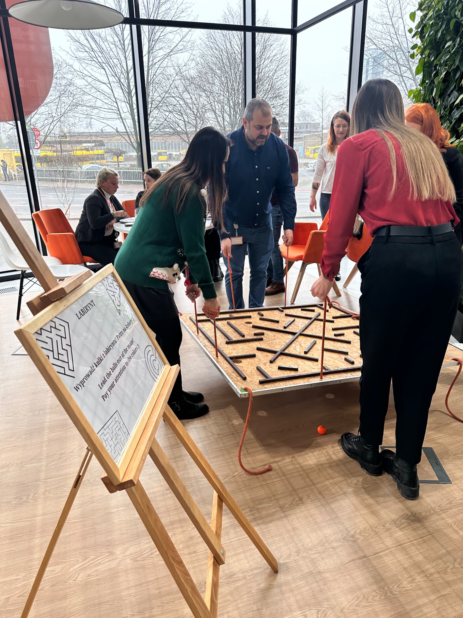 Grupa osób w biurze uczestniczy w grze zespołowej z labiryntem, używając sznurków do prowadzenia kulki; w tle okna z widokiem na miasto i inne osoby.