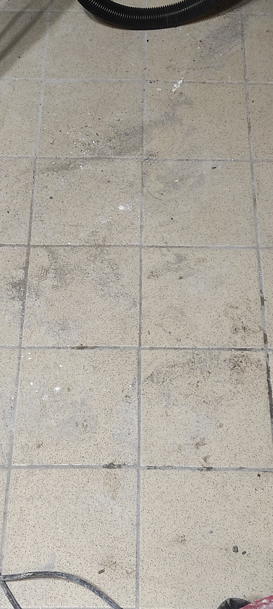 Brudna, beżowa podłoga z płytek kwadratowych, z widocznymi śladami stóp, plamami i resztkami po remoncie. Część węża od odkurzacza widoczna w górnej części kadru.