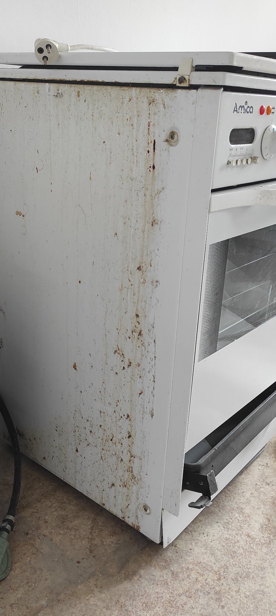 Bardzo zabrudzona, biała kuchenka Amica z widocznymi plamami i zaciekami na bocznej ściance oraz kablami zasilającymi.