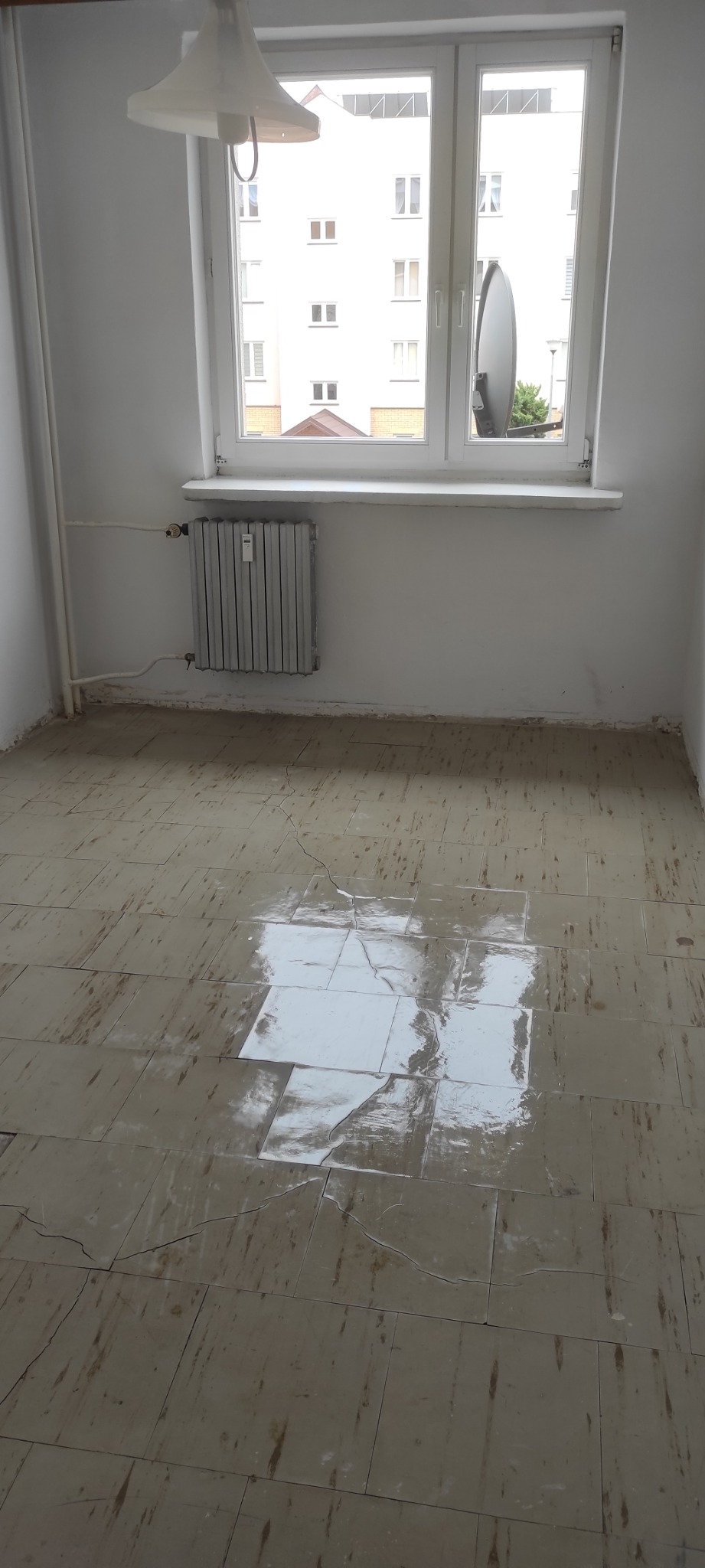 Pokój z brudną, popękaną podłogą z płytek, grzejnikiem pod oknem i lampą wiszącą u góry.