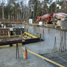 Wylany fundament z wystającymi prętami zbrojeniowymi i szalunkami w tle, z betoniarką na posesji i lasem w tle.