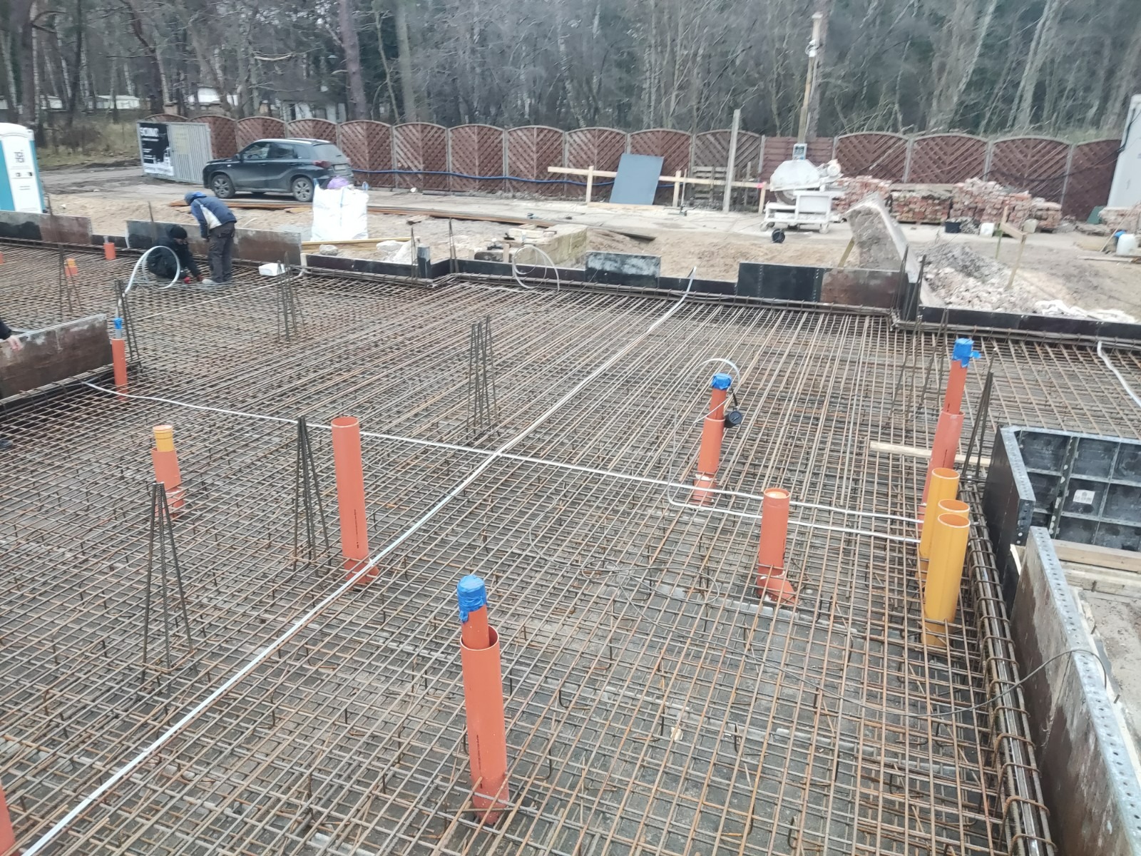 Zbrojenie fundamentów z wystającymi pionowymi rurami instalacyjnymi i pracownikiem w tle, widoczne szalunki.