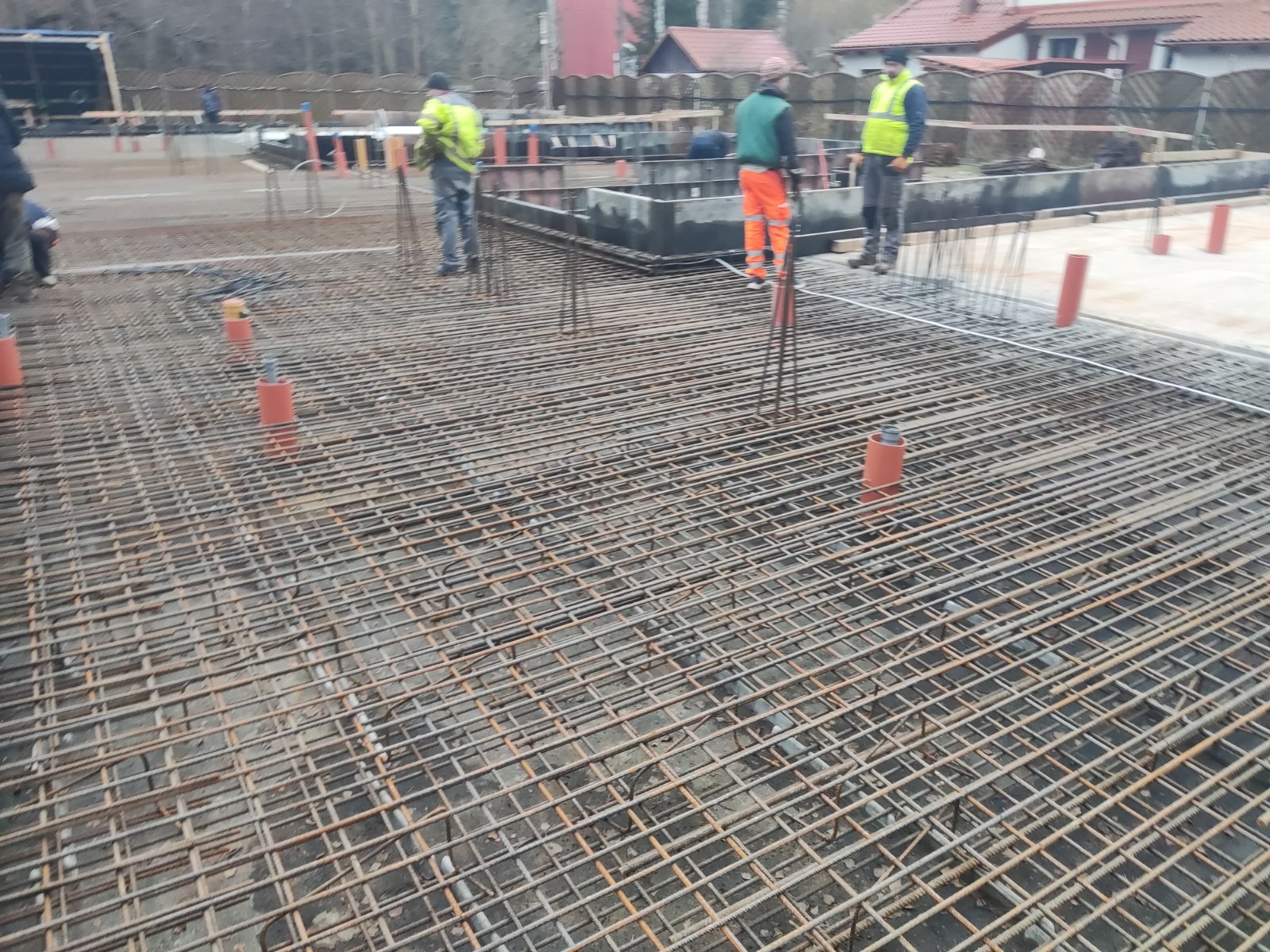 Siatka zbrojeniowa ułożona na przygotowanym terenie pod budowę z widocznymi pracownikami w odblaskowych kamizelkach i elementami szalunku w tle.
