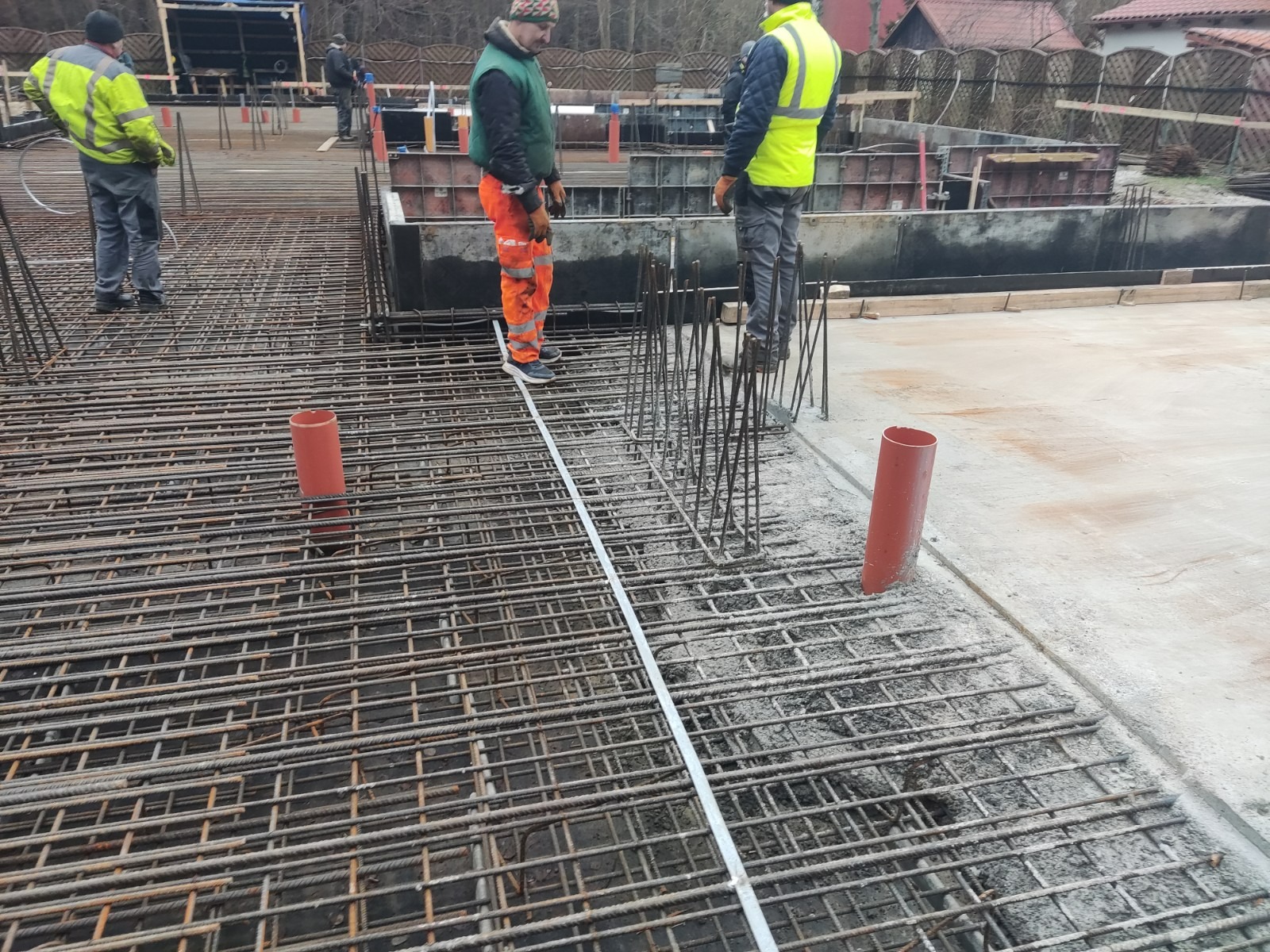 Zbrojenie fundamentów w trakcie budowy, widoczni pracownicy w odblaskowych ubraniach, szalunki i wystające pręty zbrojeniowe.