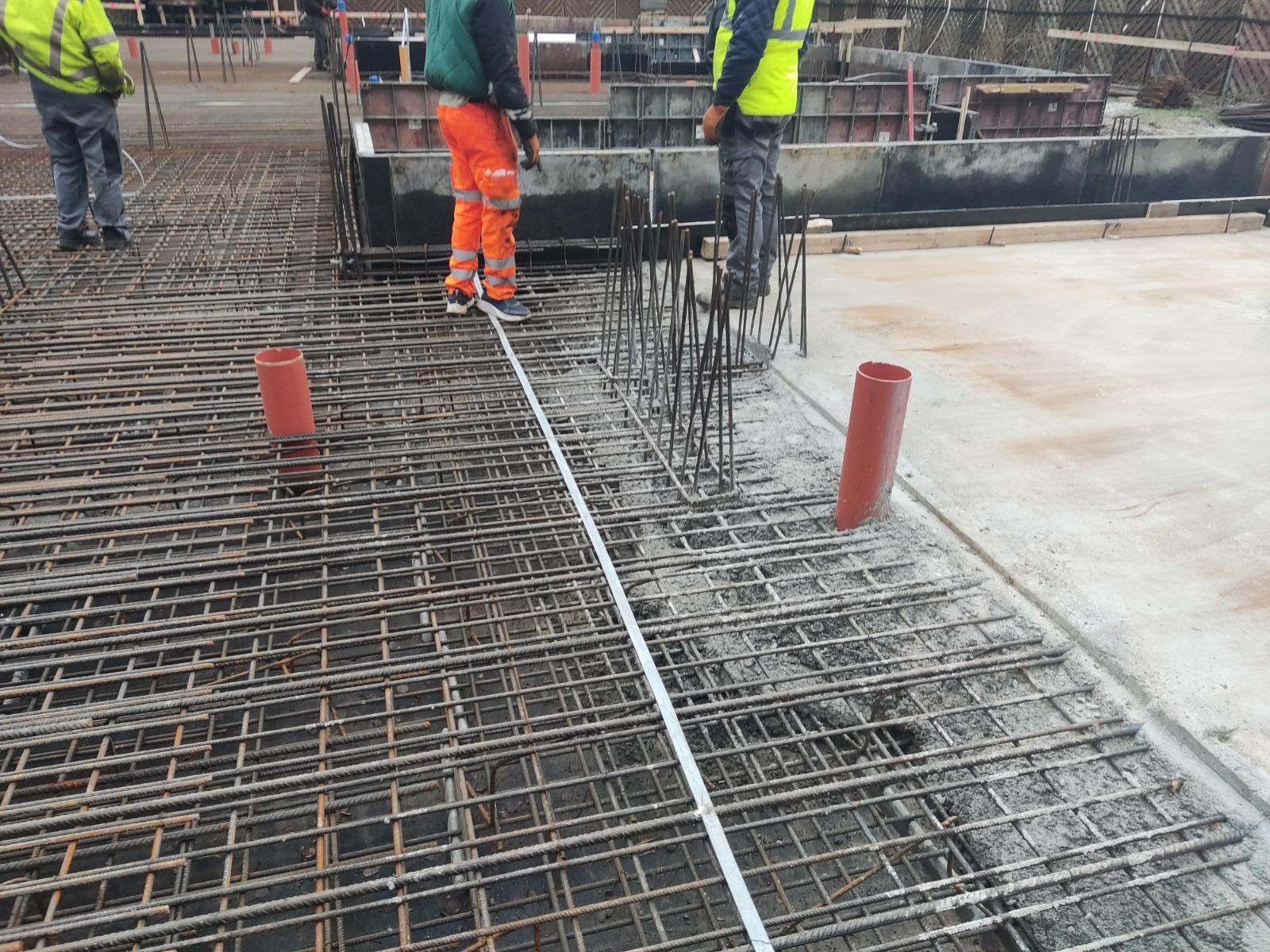 Zbrojenie fundamentów domu jednorodzinnego z widocznymi prętami stalowymi, rurami PCV i pracownikami w odzieży roboczej na placu budowy.