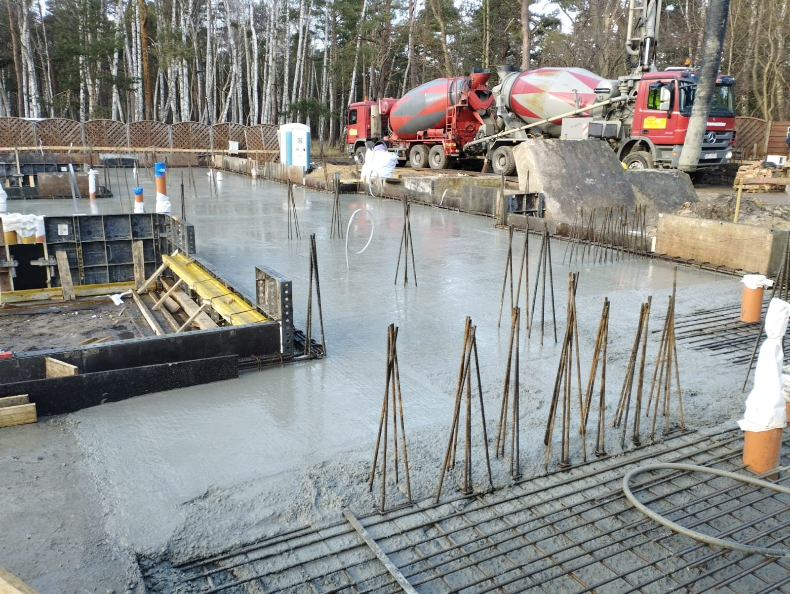 Wylany fundament z wystającymi prętami zbrojeniowymi; w tle dwa betonowozy na tle lasu.