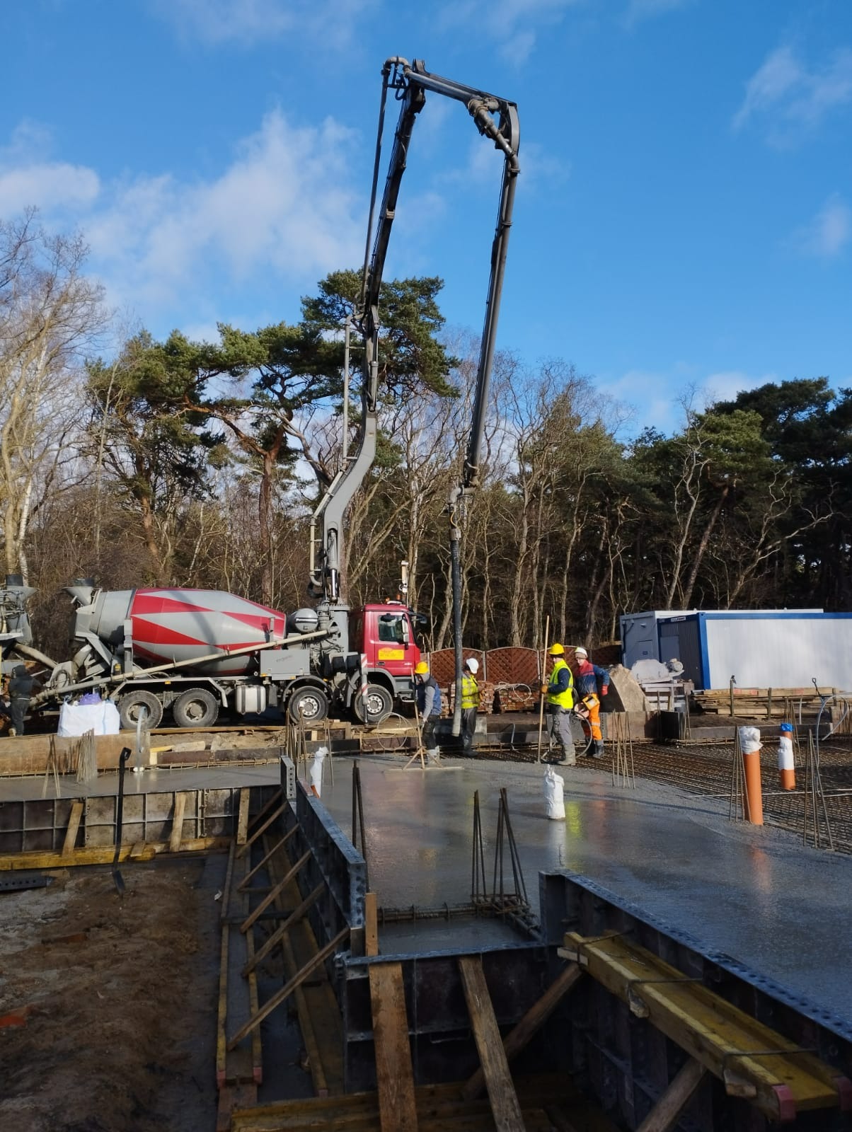 Wylewanie betonu na fundamenty domu jednorodzinnego za pomocą pompy do betonu, widoczni pracownicy w kaskach i kamizelkach odblaskowych, szalunki i zbrojenie w przygotowanym wykopie.