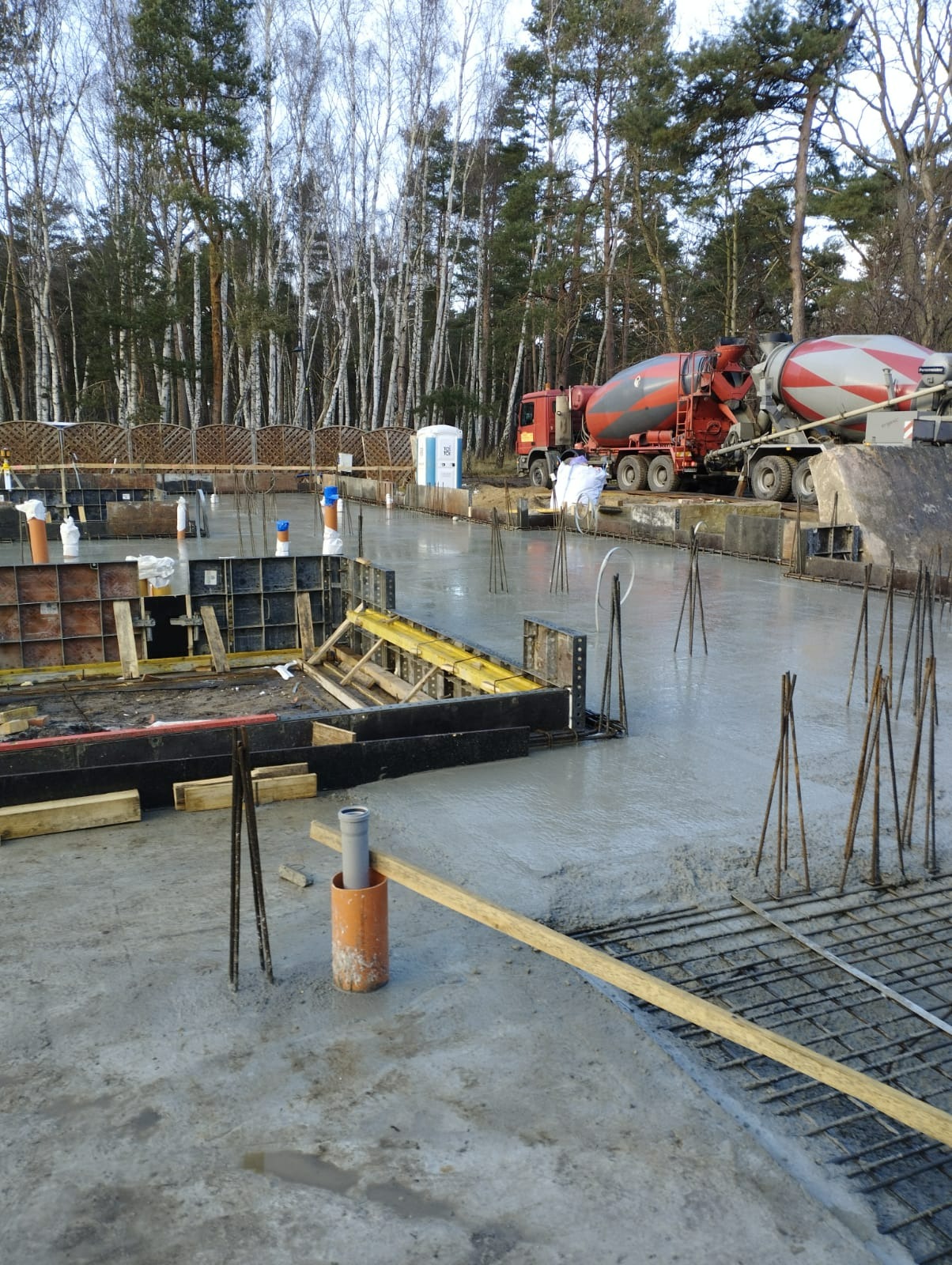 Wylany fundament z wystającymi prętami zbrojeniowymi i szalunkami w tle, z betoniarką na posesji i lasem w tle.