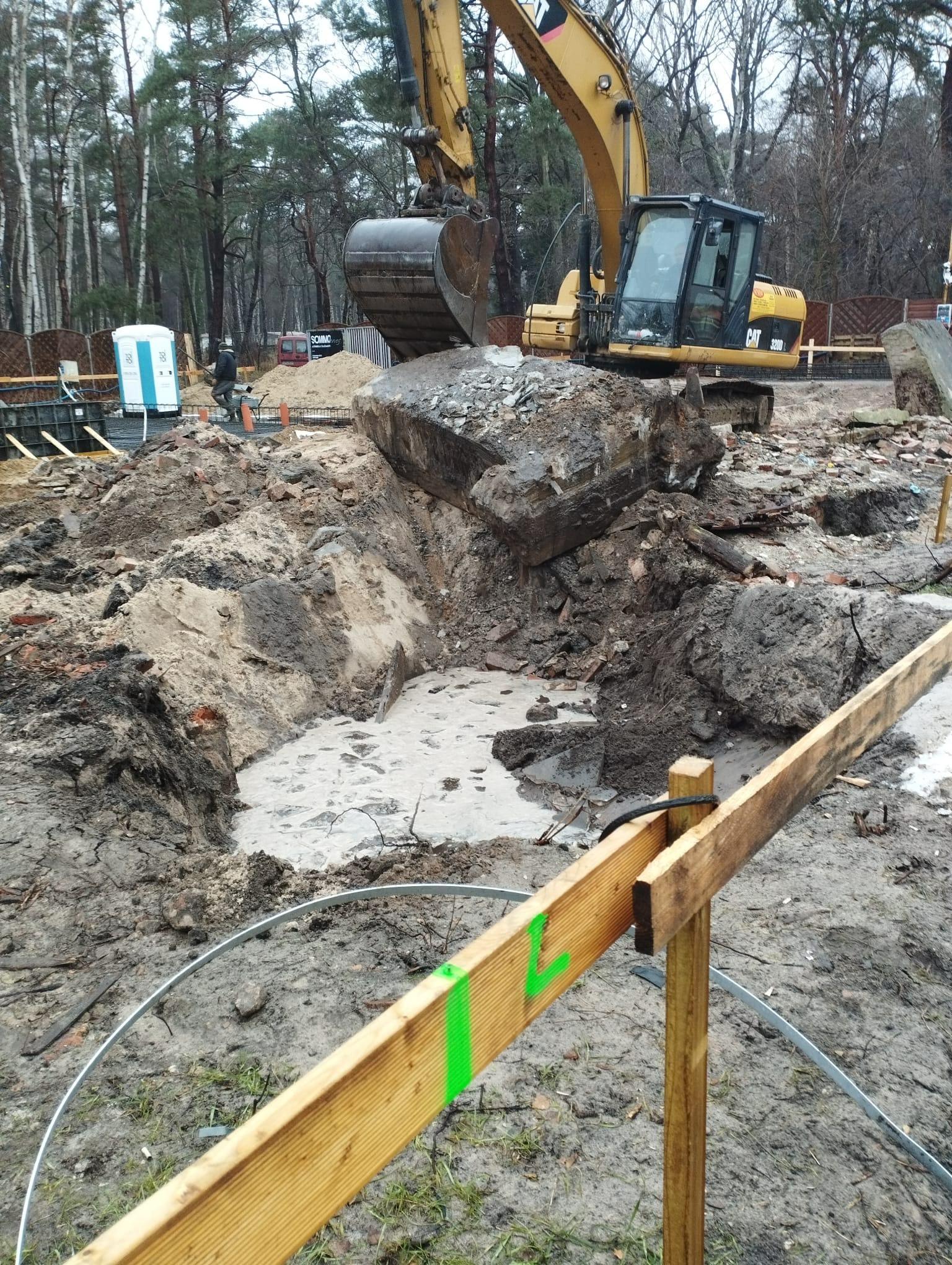 Żółta koparka CAT umieszcza duży kamień w wykopie wypełnionym wodą, widoczne szalunki fundamentów i przenośna toaleta w tle.