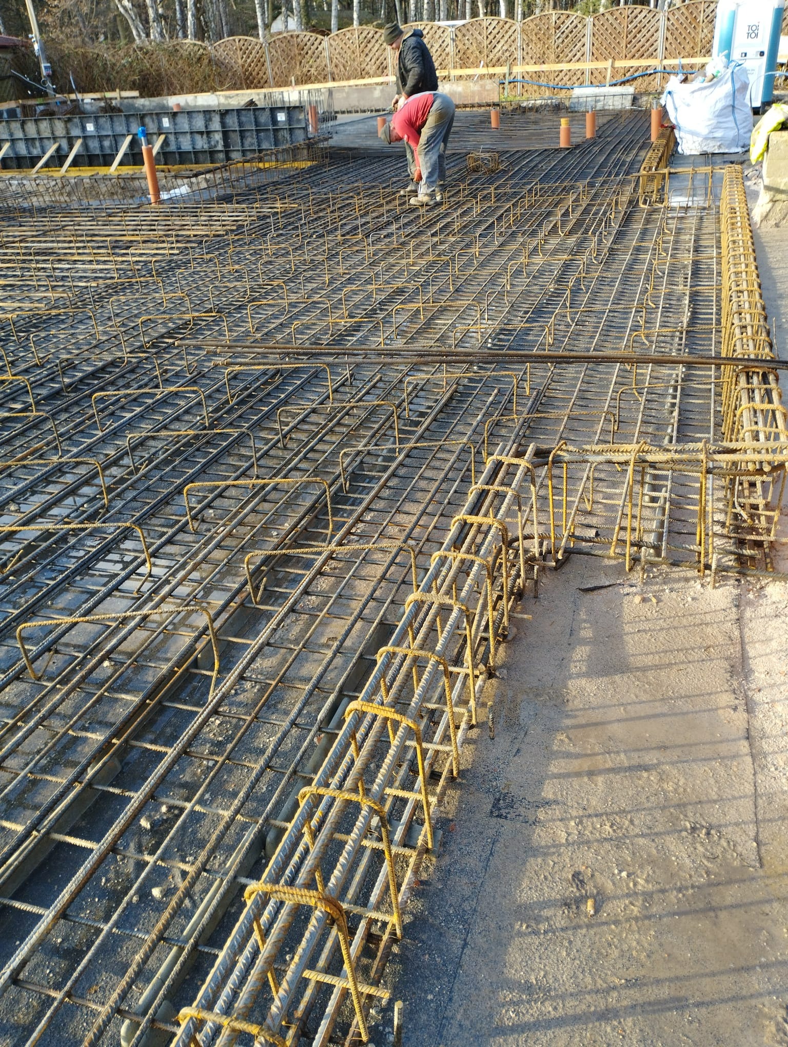 Zbrojenie fundamentów domu jednorodzinnego, widoczne stalowe pręty i siatki zbrojeniowe, dwóch pracowników w tle.