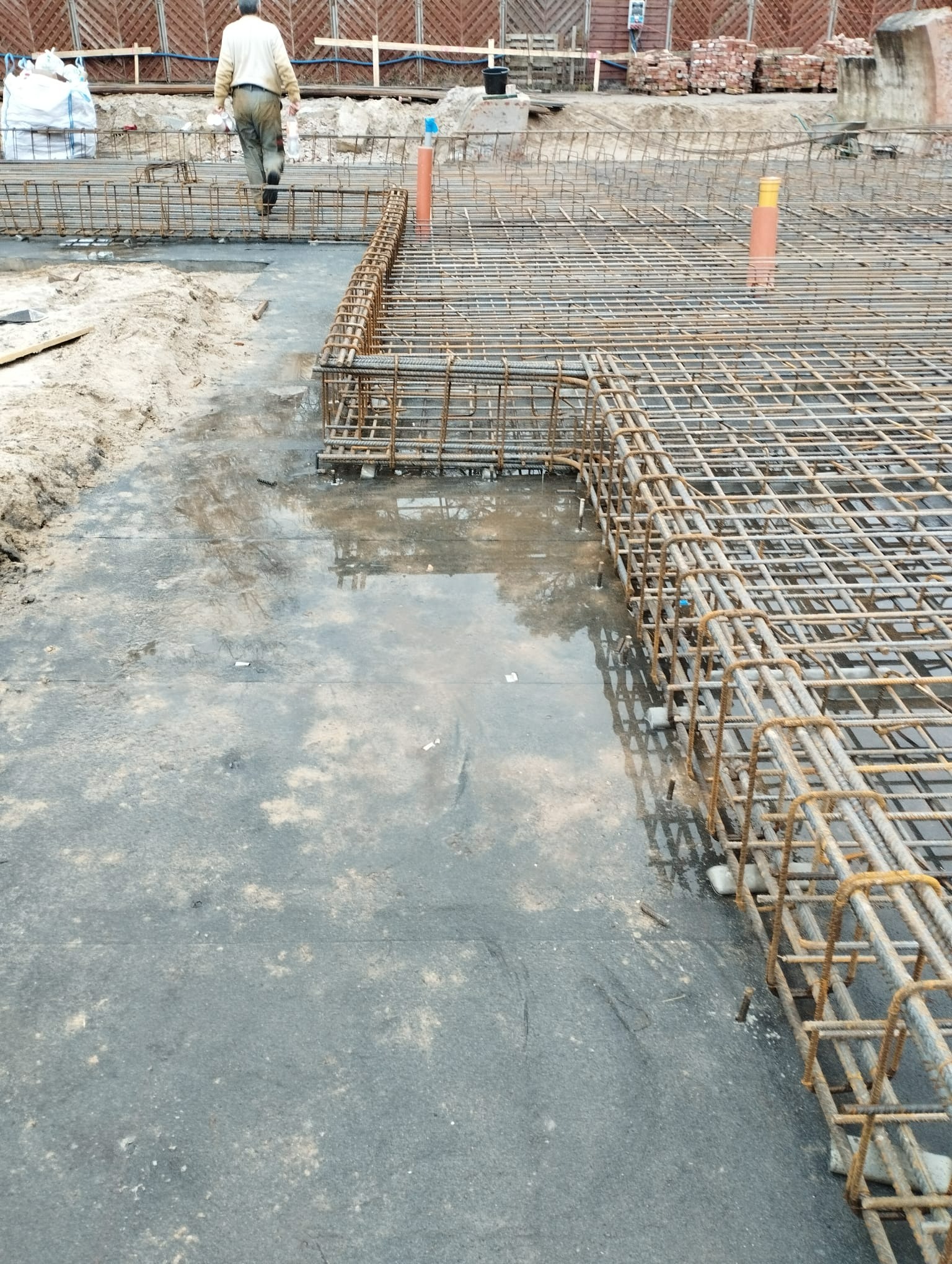 Zbrojenie fundamentów domu w trakcie budowy, widoczne stalowe pręty połączone w siatkę, na mokrym podłożu, w tle cegły i pracownik budowlany.