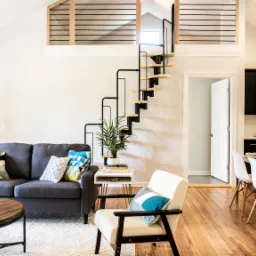 Jasne wnętrze domu z antresolą, spiralnymi schodami, szarą sofą i otwartą kuchnią z czarnymi szafkami.