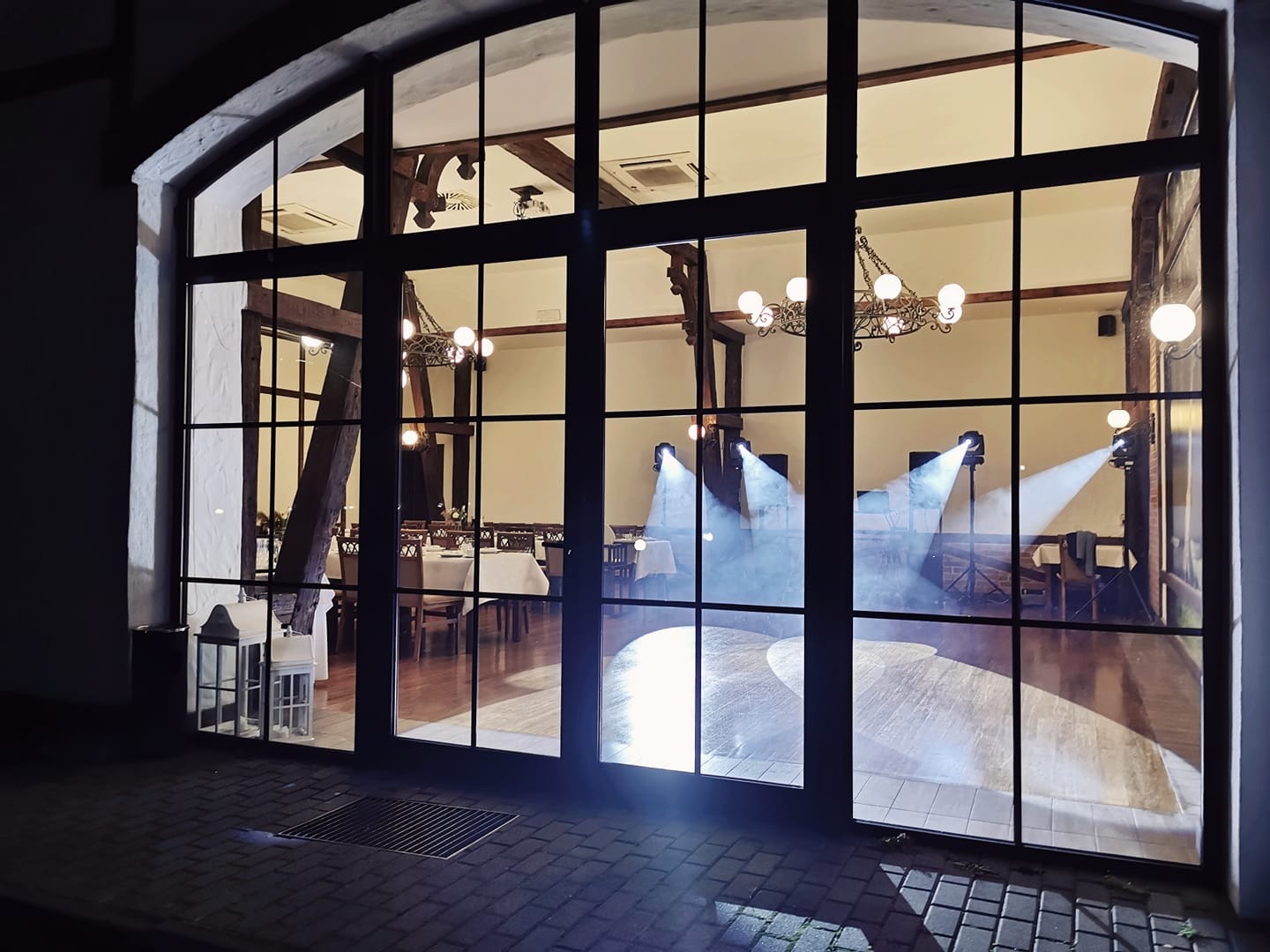 Sala balowa z parkietem, oświetlona reflektorami i wiszącymi żyrandolami, widoczna przez duże okno z czarnymi szprosami, ustawione stoły z białymi obrusami.