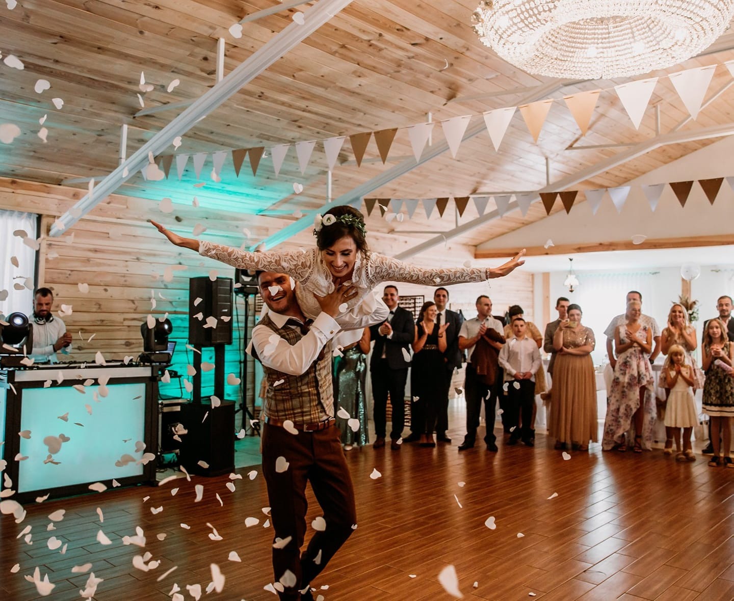 Pan młody trzyma pannę młodą na rękach podczas pierwszego tańca, konfetti w kształcie serc w powietrzu, w tle DJ, goście weselni i dekoracje sali.