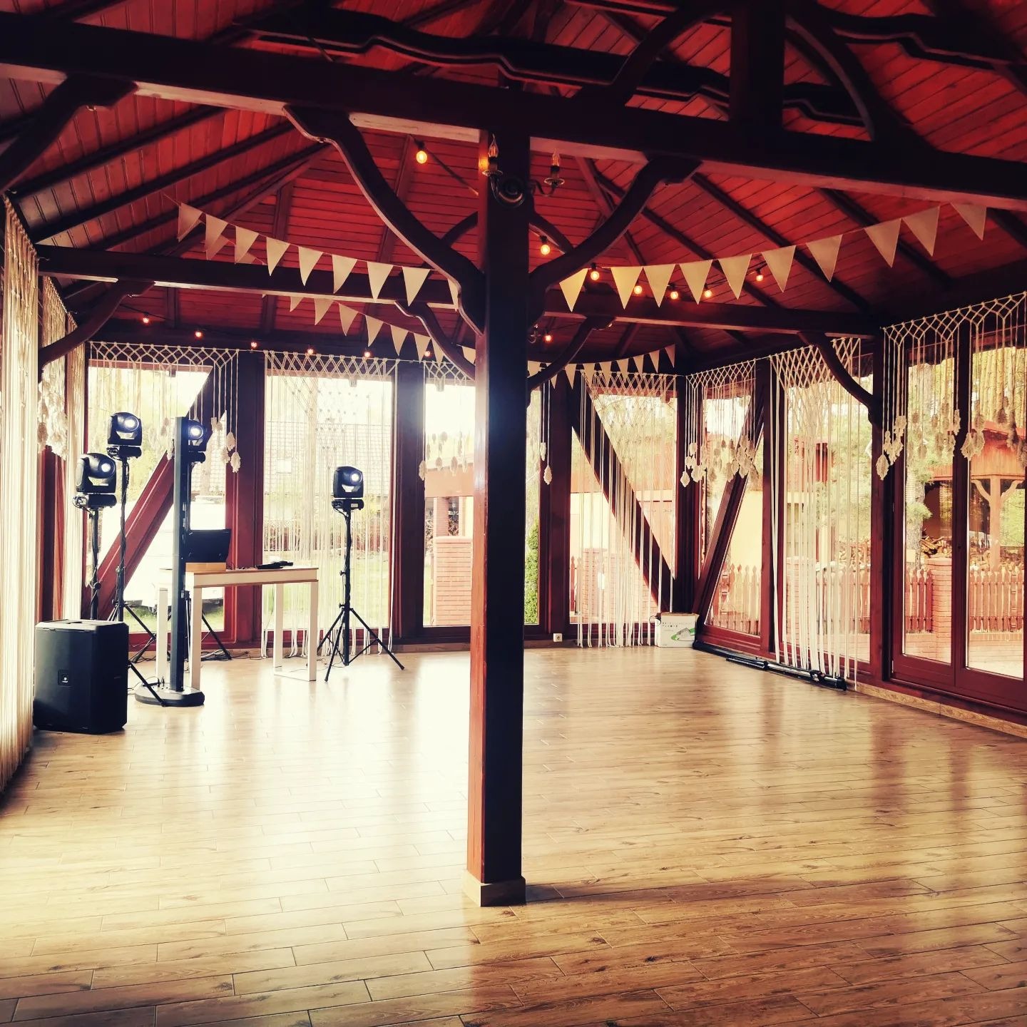 Przygotowane do imprezy drewniane wnętrze altany z nagłośnieniem, oświetleniem i dekoracjami w stylu rustykalnym, w tym girlandy i makramy przy oknach.