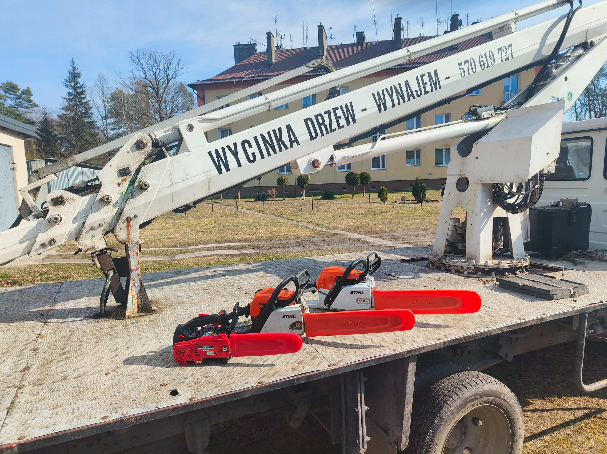 Platforma samochodu z podnośnikiem koszowym, na której leżą trzy piły spalinowe marki Stihl z czerwonymi osłonami na prowadnicach, w tle budynek mieszkalny i błękitne niebo.
