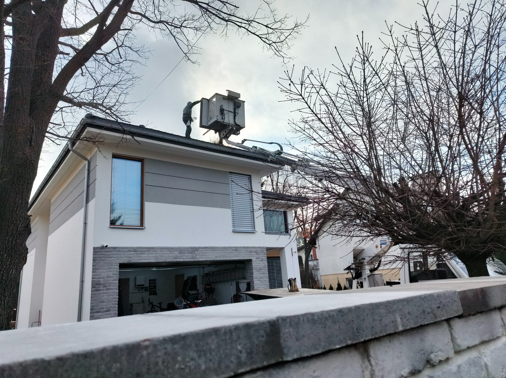 Pracownik w podnośniku koszowym na dachu dwukondygnacyjnego domu, wykonujący prace na wysokości, widok zza murku.