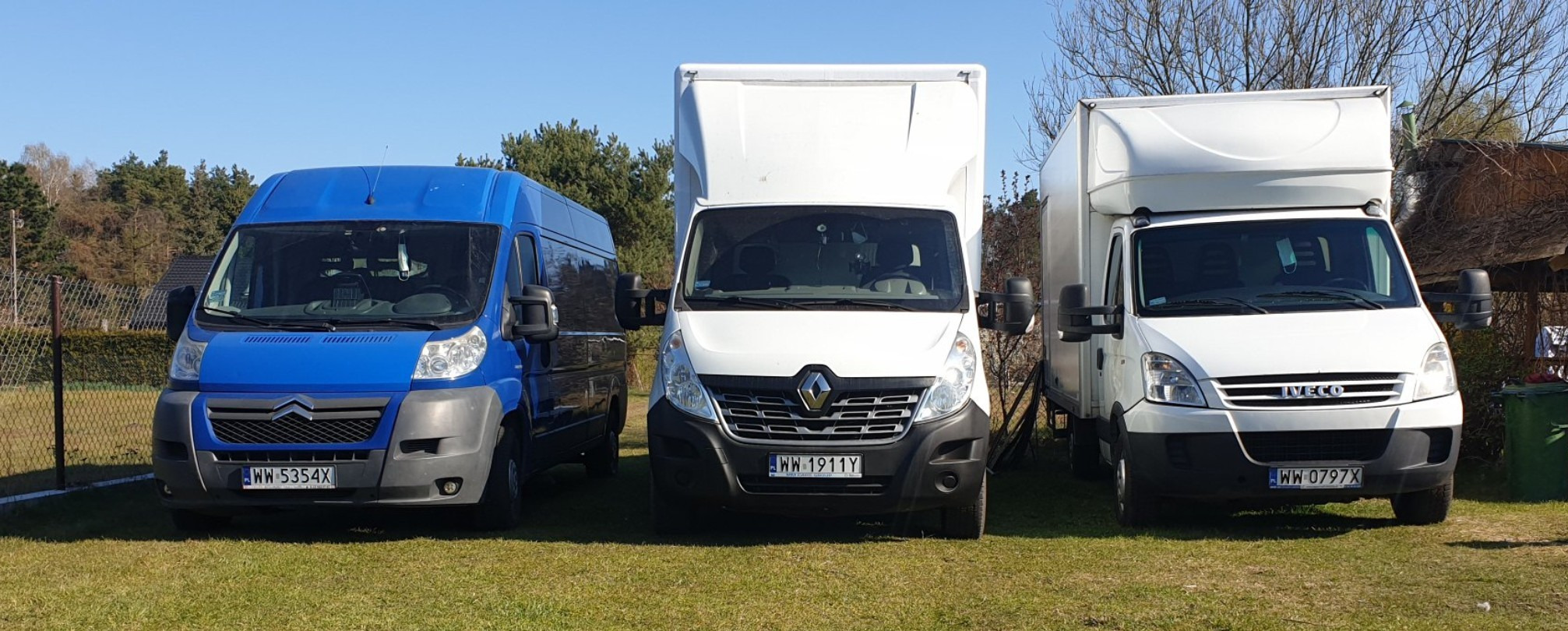 Trzy busy transportowe różnych marek (Citroen, Renault, Iveco) stojące obok siebie na trawiastym terenie w słoneczny dzień.