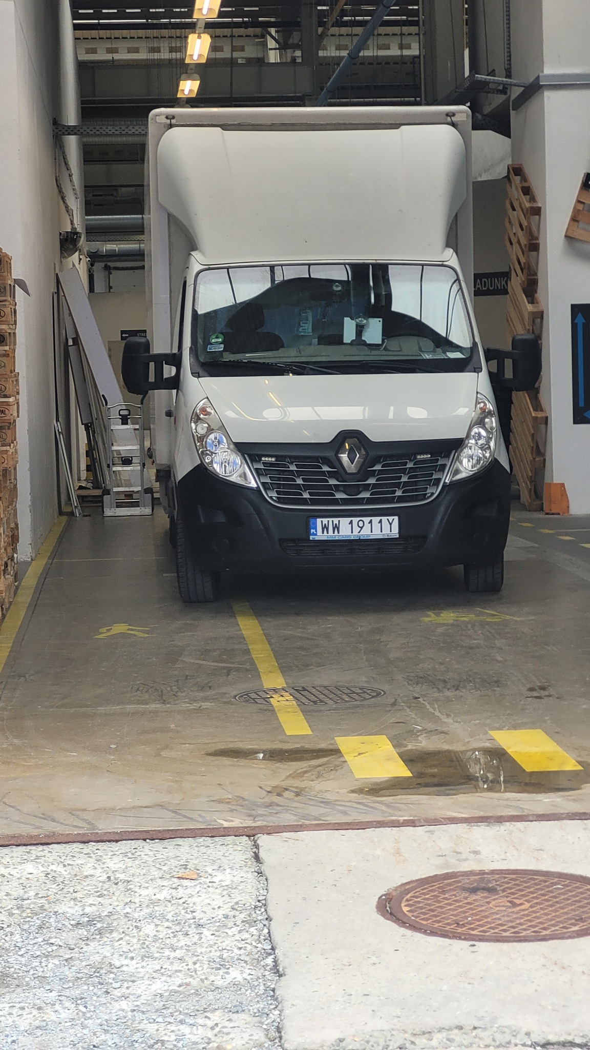 Biały Renault Master z wysoką zabudową zaparkowany w hali magazynowej, widoczne żółte pasy na posadzce i regały w tle.