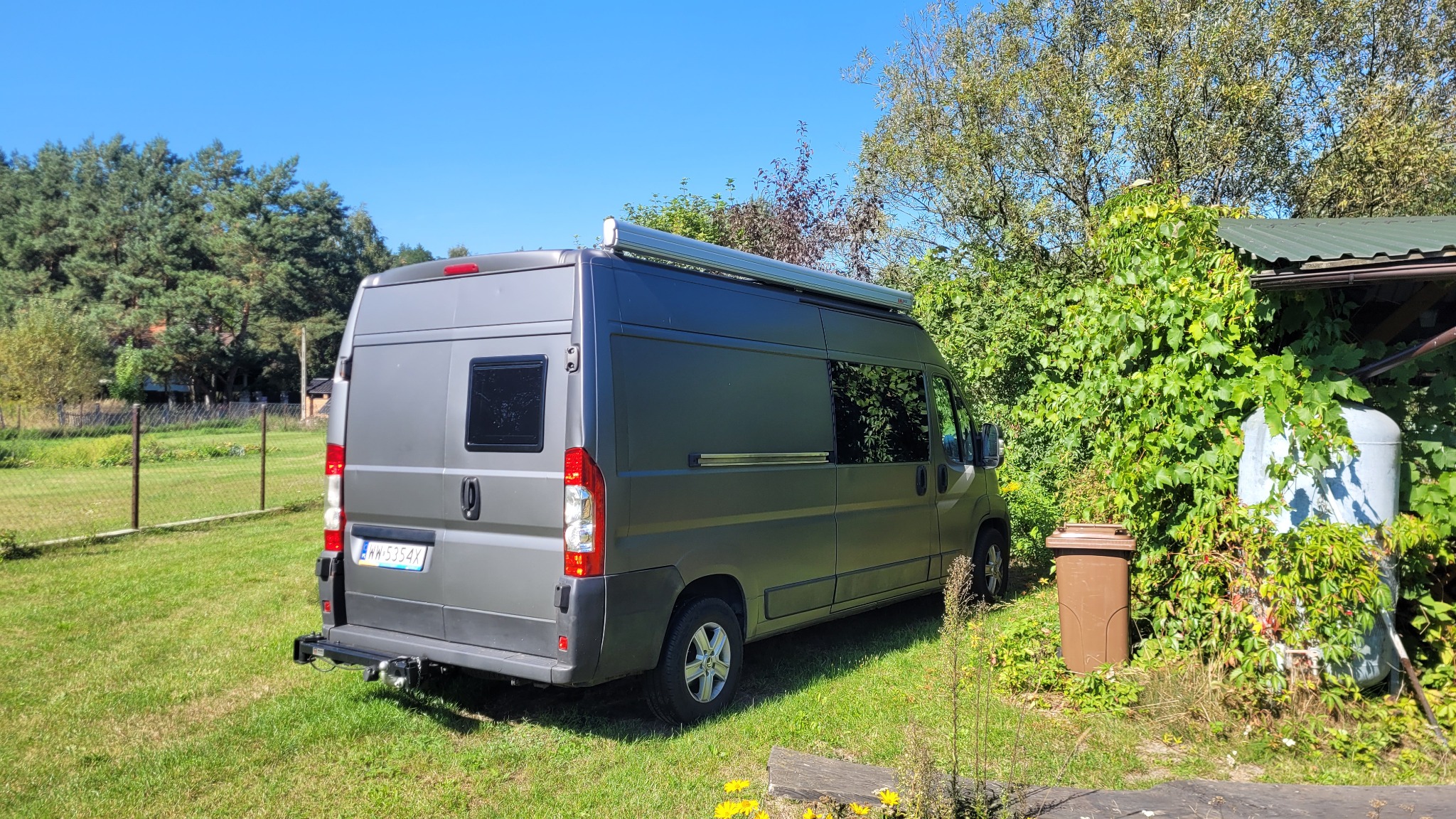 Szary kamper z wysuwaną markizą zaparkowany na trawniku w słoneczny dzień, widok z tyłu.