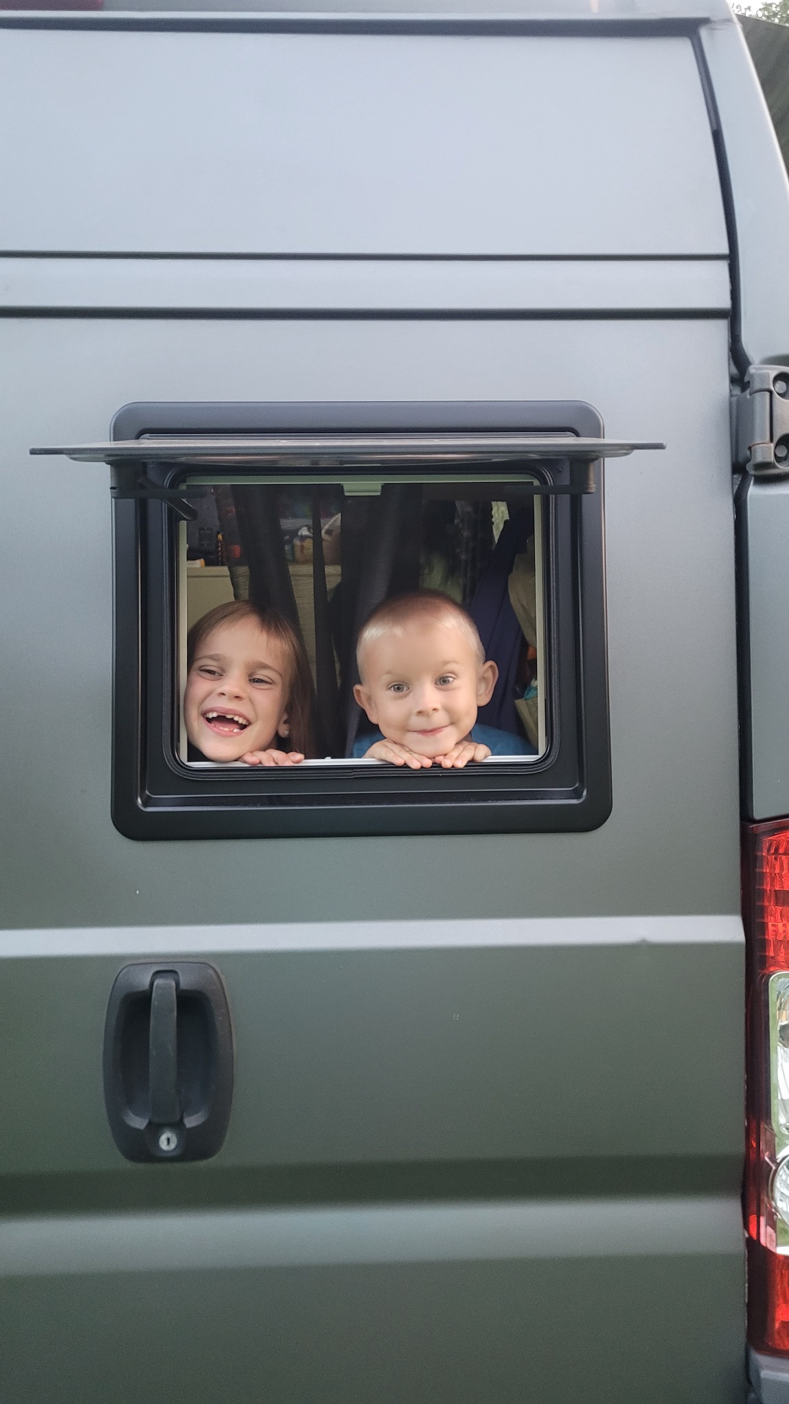 Dwoje dzieci, dziewczynka z szerokim uśmiechem i mały chłopiec, wyglądają przez otwarte okno w szarym busie typu kamper.