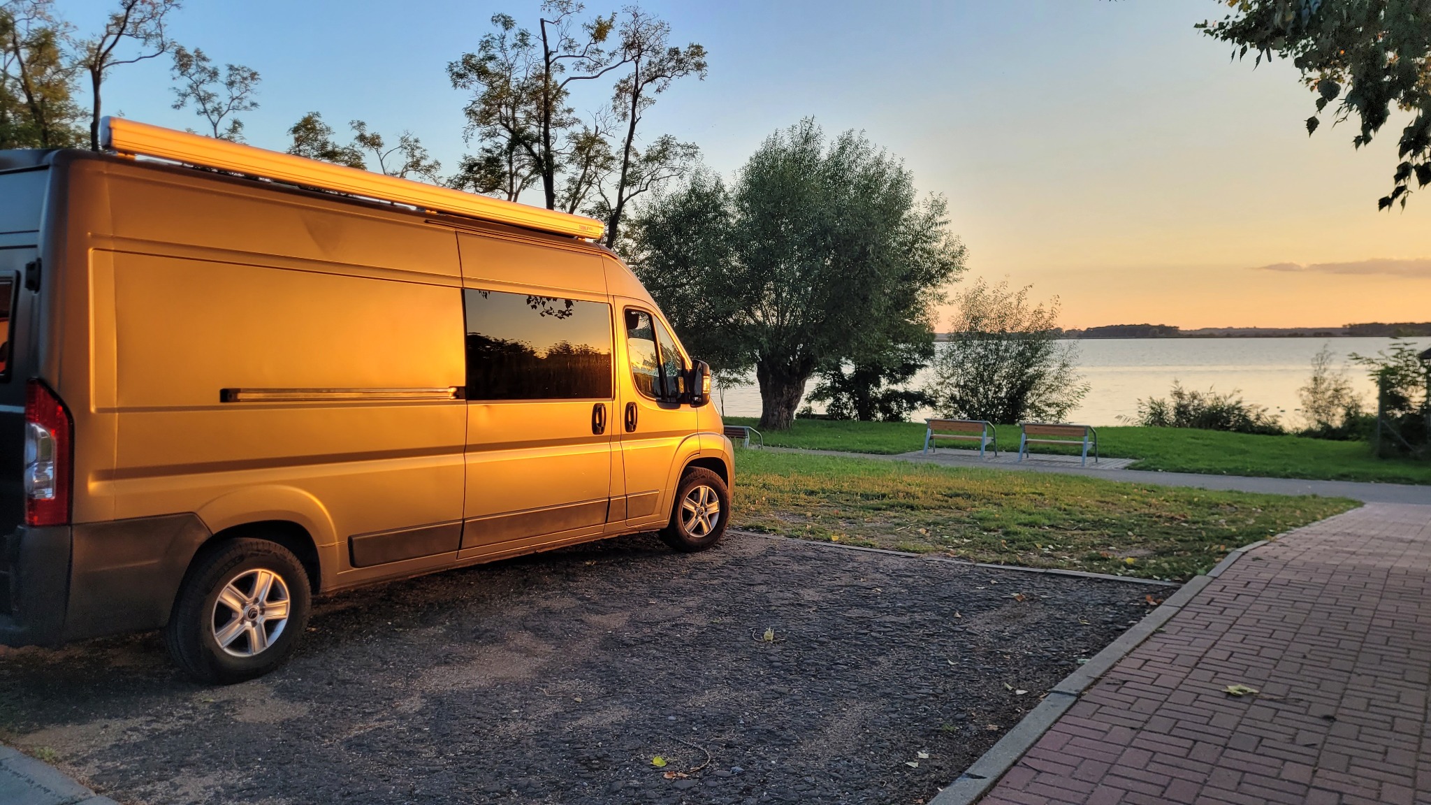 Złoty kamper z rozłożoną markizą parkuje nad jeziorem o zachodzie słońca, widoczne dwie ławki na trawie.