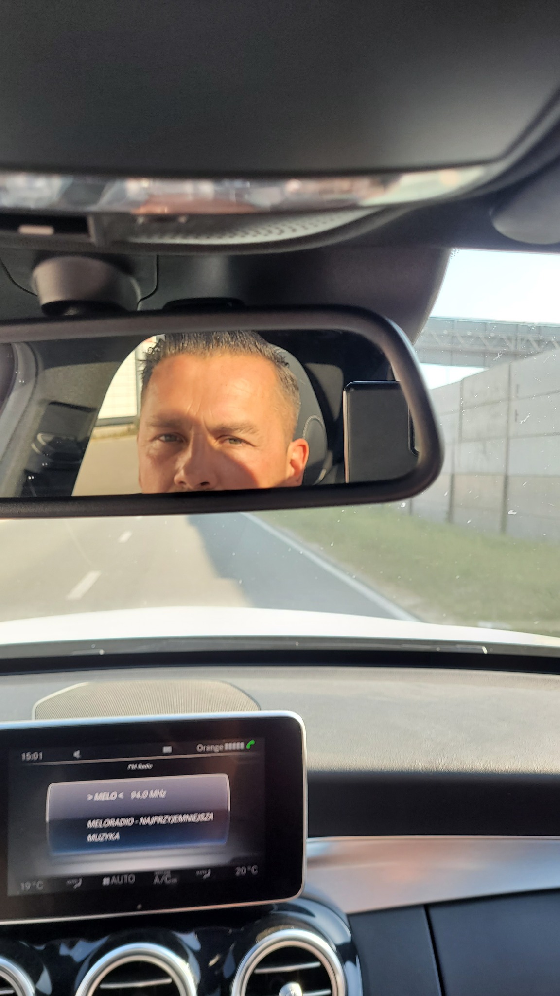 Kierowca widziany w lusterku wstecznym samochodu marki Mercedes, z widocznym radiem ustawionym na stację Meloradio.