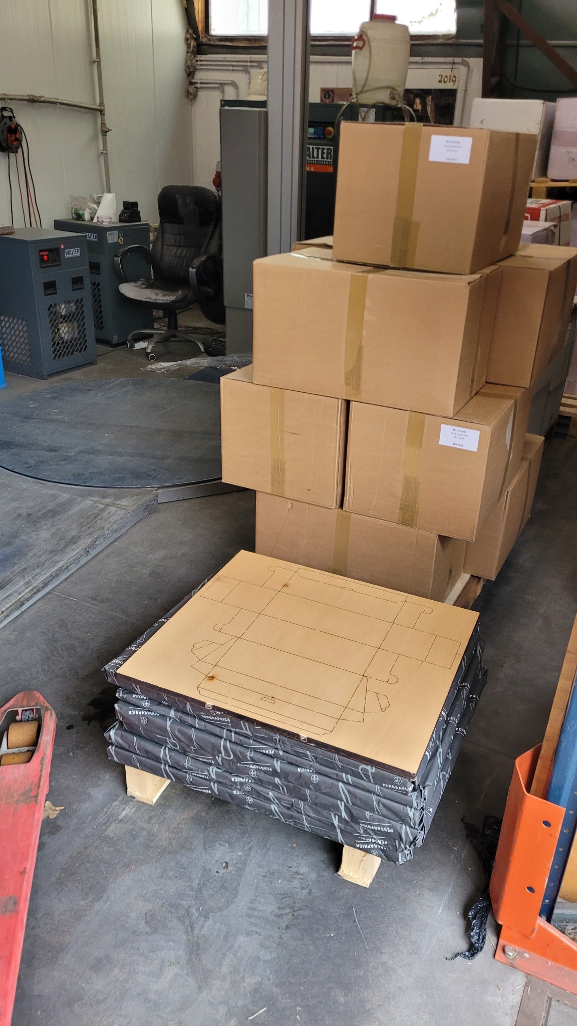 Stos zapakowanych arkuszy z wyciętym szablonem kartonowego pudełka, ułożonych na palecie w hali magazynowej, w tle widoczne kartony i sprzęt przemysłowy.