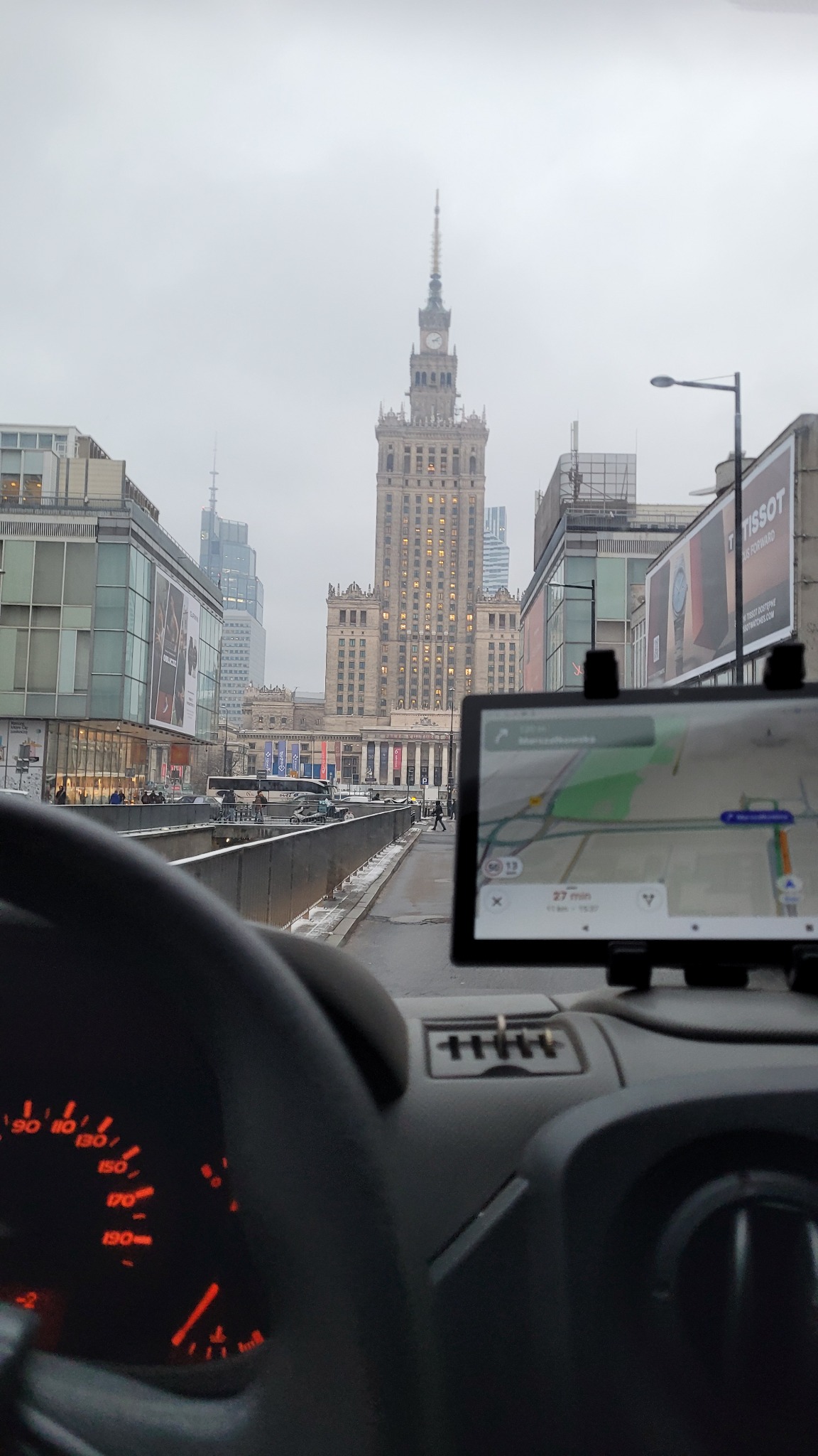 Widok z wnętrza pojazdu na Pałac Kultury i Nauki w Warszawie, nawigacja GPS na tablecie pokazuje trasę, szaro-bura pogoda.