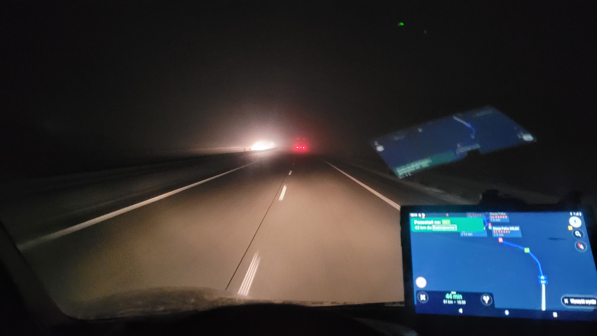 Jazda nocą autostradą w gęstej mgle, widok z wnętrza pojazdu na drogę oświetloną światłami nadjeżdżających aut, nawigacja GPS wyświetla trasę.