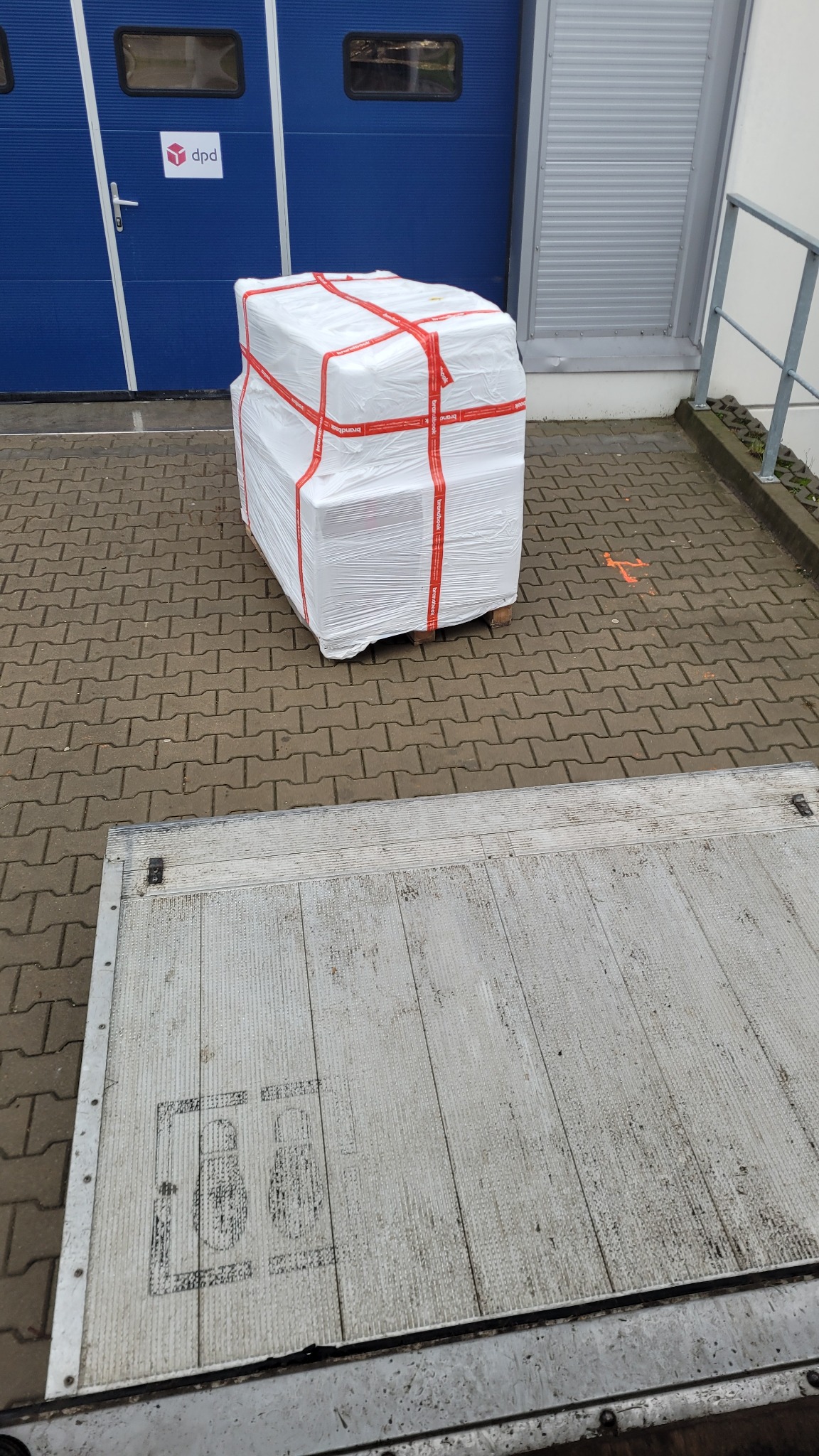 Paczka owinięta folią i zabezpieczona taśmami na palecie, częściowo widoczna platforma załadunkowa samochodu dostawczego, w tle brama magazynowa DPD.