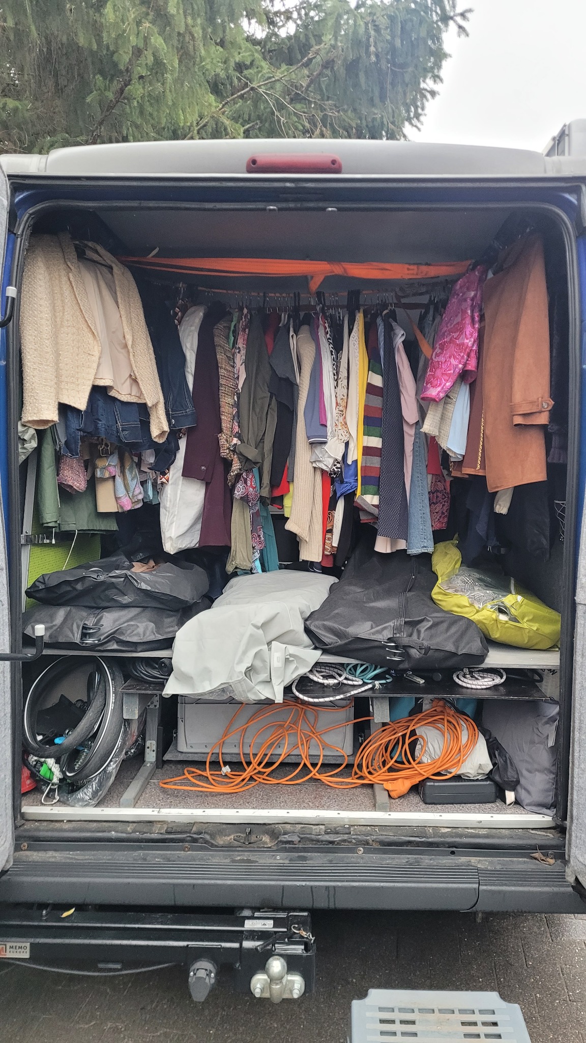 Wnętrze busa zaadaptowane jako mobilna garderoba, ubrania wiszące na wieszakach, torby i akcesoria na półkach, pomarańczowe kable zwinięte na dole.