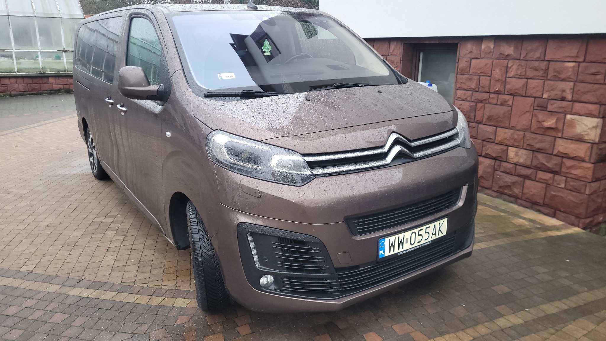 Brązowy Citroen SpaceTourer zaparkowany na kostce brukowej w deszczowy dzień, widoczny z przodu, z zielonym odświeżaczem powietrza w kształcie choinki wiszącym na lusterku.