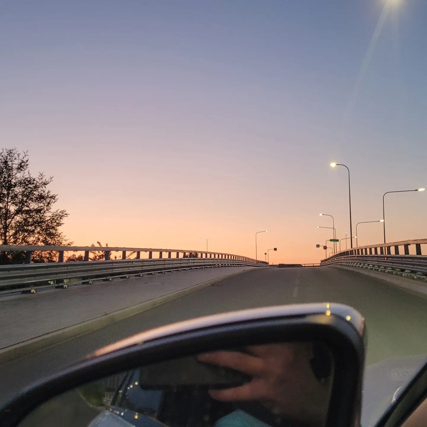 Widok z wnętrza samochodu na drogę wznoszącą się na wiadukcie o zmierzchu, odbicie w lusterku bocznym.