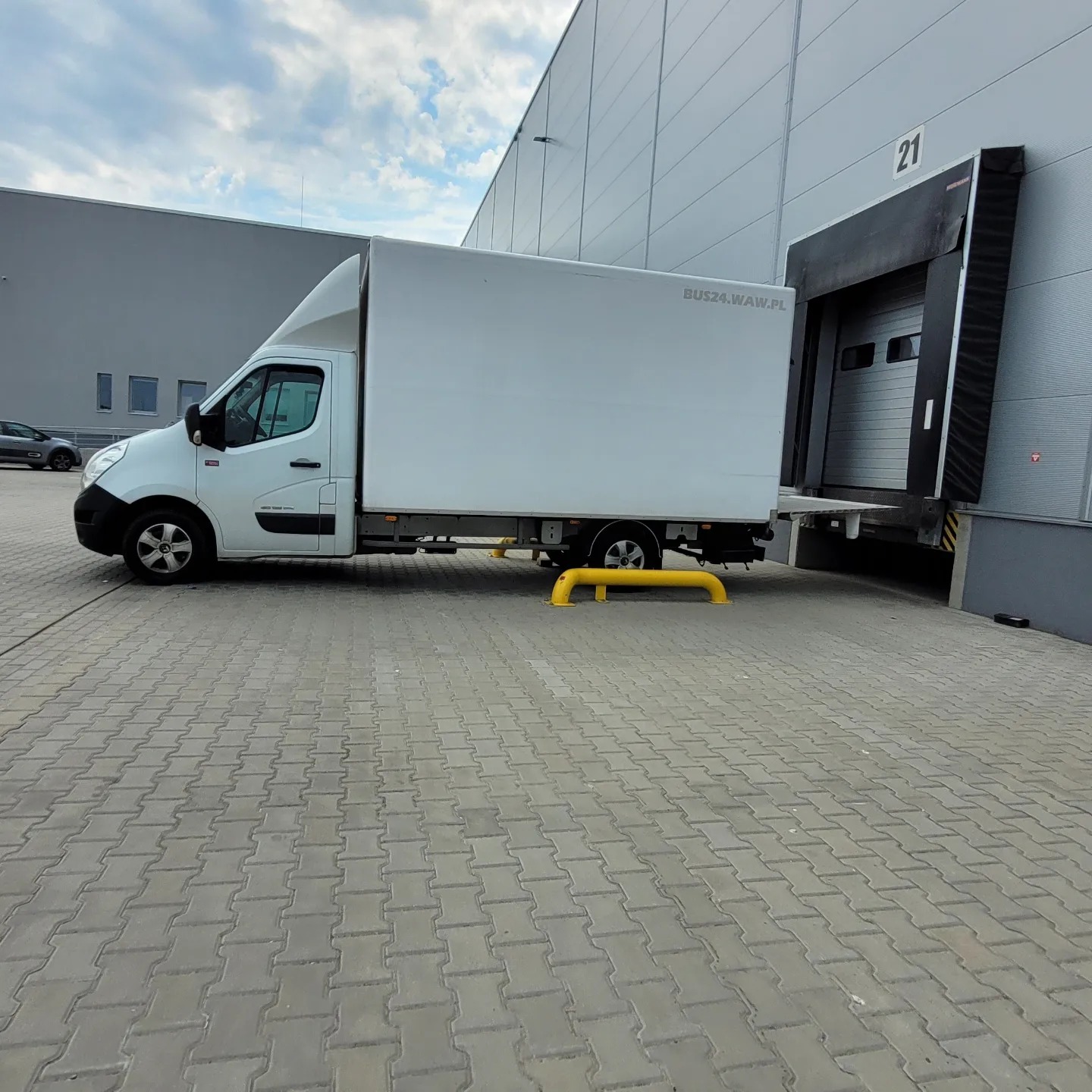 Biały furgon dostawczy zaparkowany przy rampie załadunkowej magazynu, z widocznym adresem strony internetowej na boku.