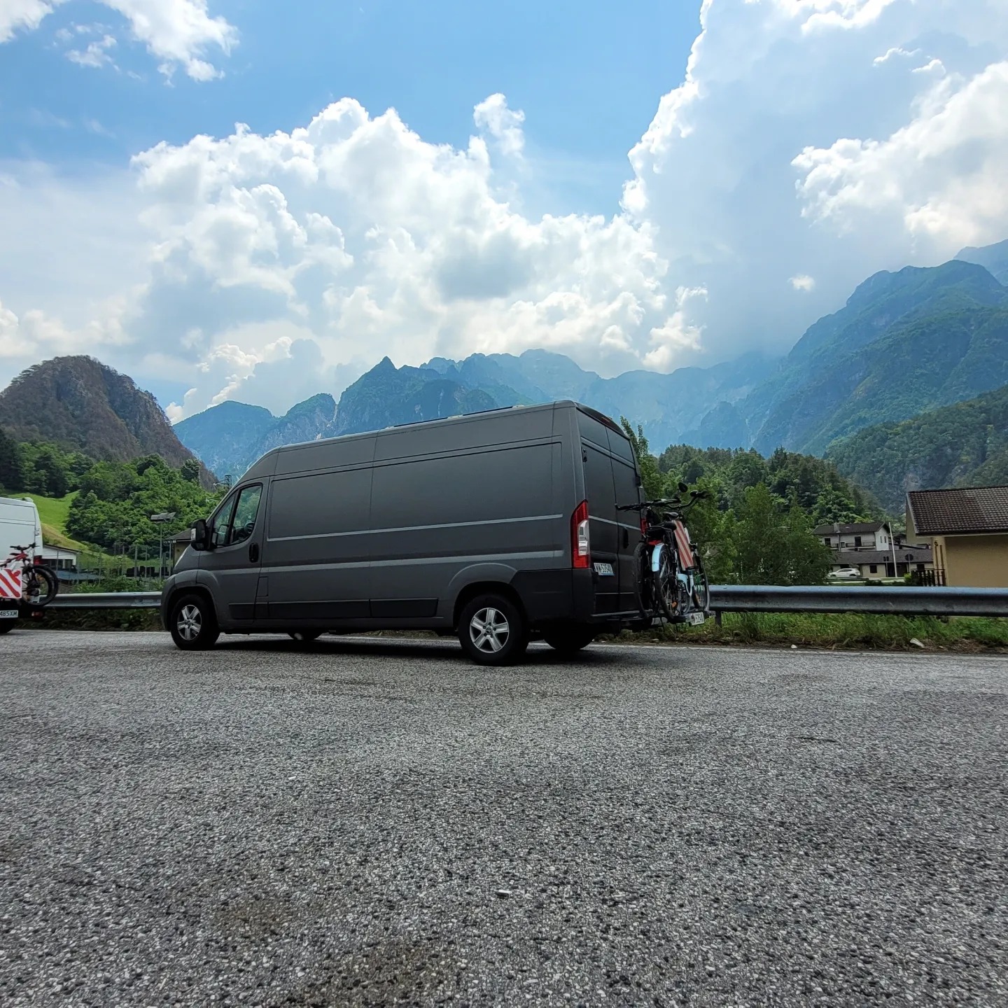 Szary bus z rowerami na bagażniku rowerowym, zaparkowany na tle górskiego krajobrazu z błękitnym niebem i obłokami.