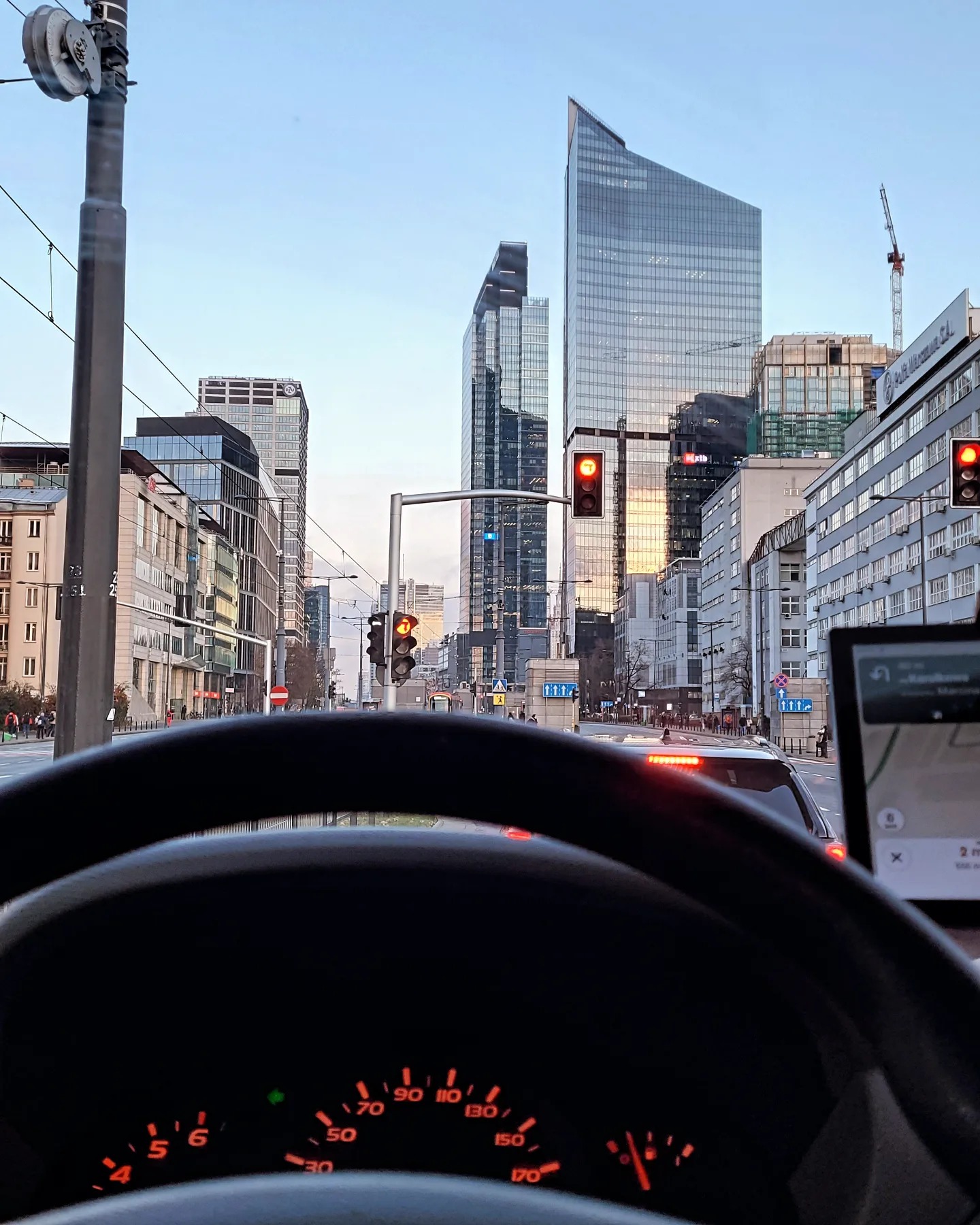 Widok z wnętrza pojazdu na miasto z wysokimi budynkami i sygnalizacją świetlną, widoczny fragment deski rozdzielczej i nawigacji GPS