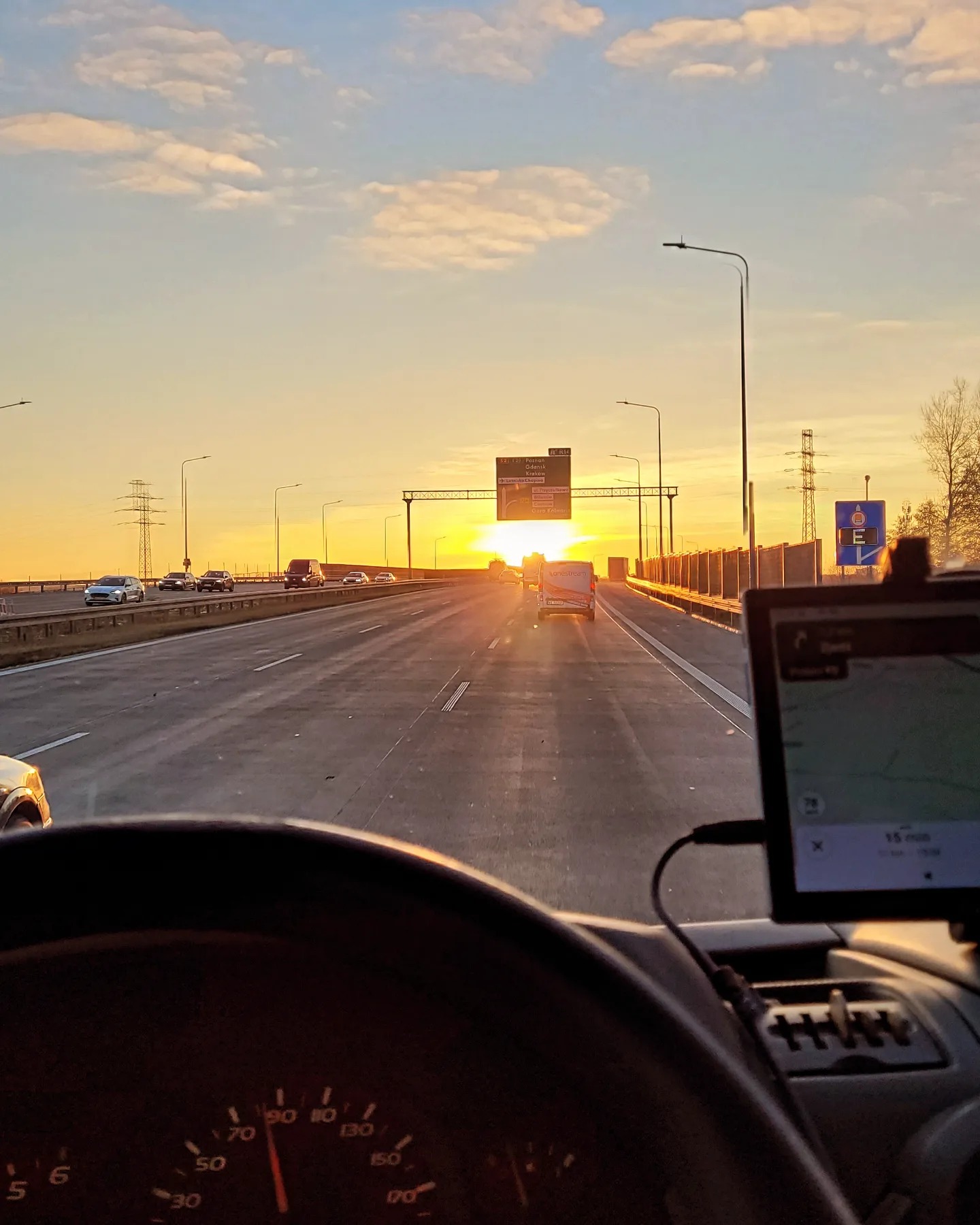 Widok z kabiny busa na autostradę o wschodzie słońca, z widocznym nawigatorem GPS i innymi pojazdami w oddali.