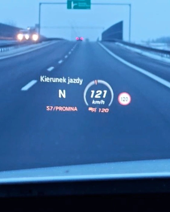 Widok z wnętrza pojazdu na drogę ekspresową S7 w kierunku Fromborka, z wyświetlaczem HUD pokazującym prędkość 121 km/h i kierunek jazdy na północ.