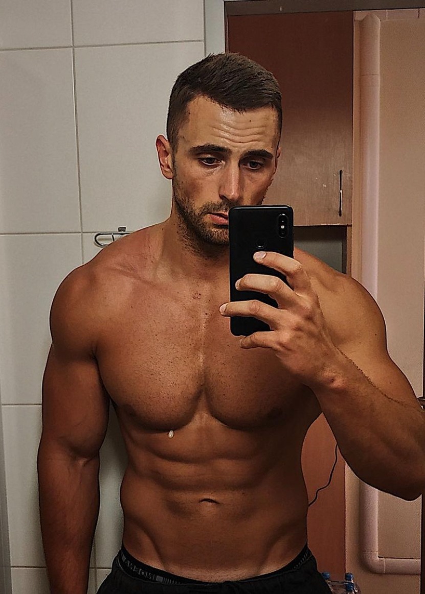 Mężczyzna robi selfie, prezentując wyrzeźbioną sylwetkę w łazience z białymi kafelkami.