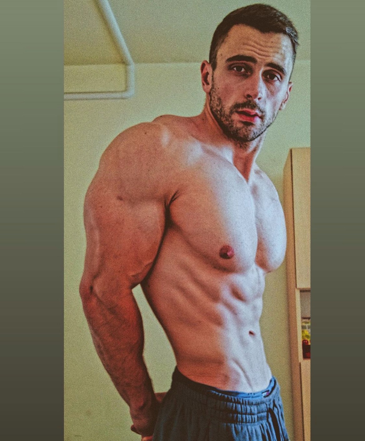 Mężczyzna z widoczną muskulaturą brzucha i ramion, pozujący w szarych spodenkach na tle ściany i szafki.