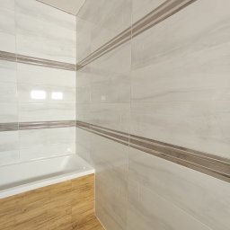 Usługi Remontowo- Budowlane MP Bud Michał Pawluszek - Wykończona łazienka z białymi płytkami imitującymi marmur, z poziomymi paskami w kolorze taupe, białą wanną i podłogą z paneli imitujących drewno.