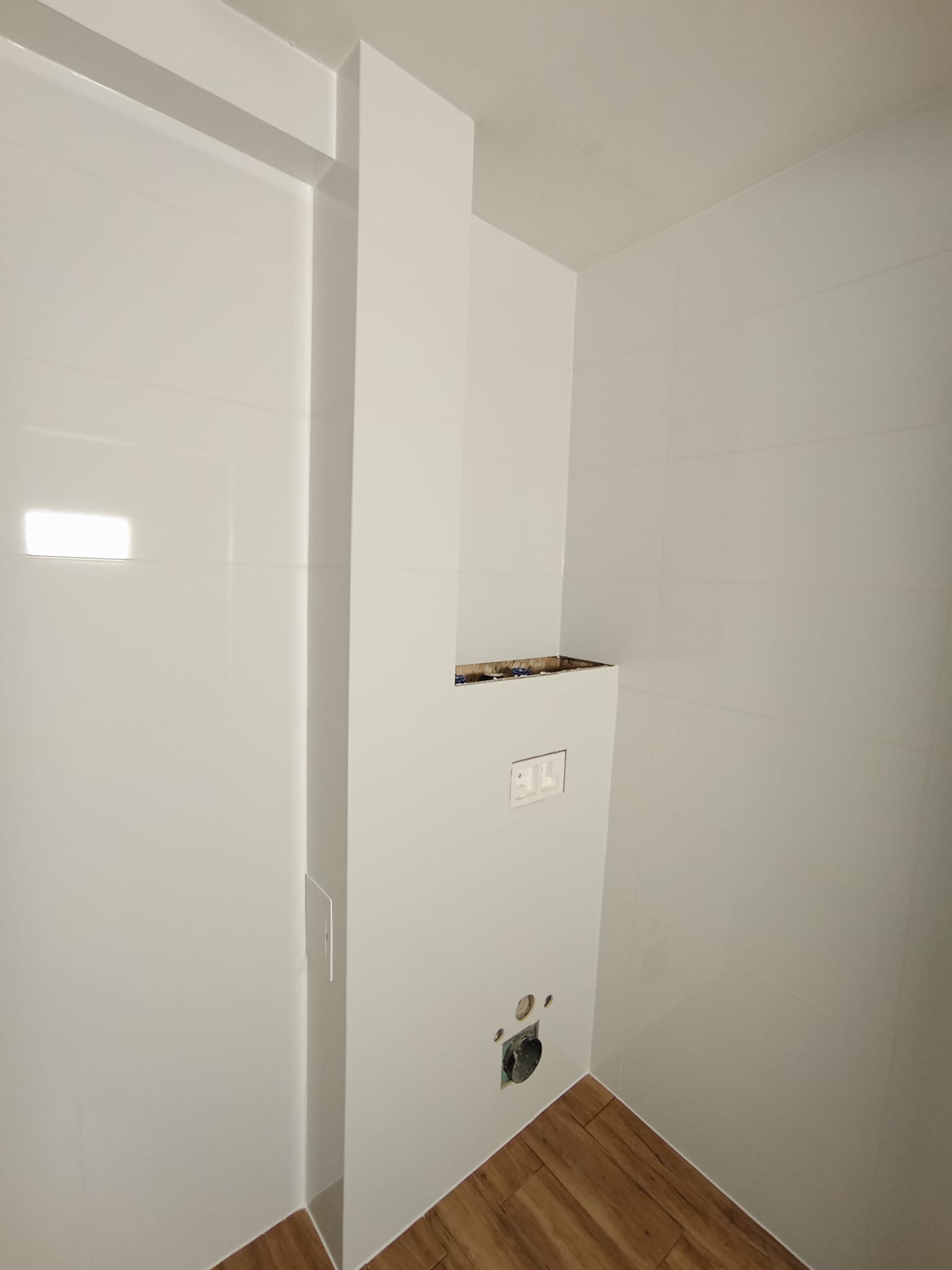 Fragment łazienki w trakcie remontu: białe, błyszczące płytki ścienne, drewnopodobne panele podłogowe, zabudowa podtynkowego stelaża WC z widoczną instalacją elektryczną i hydrauliczną.