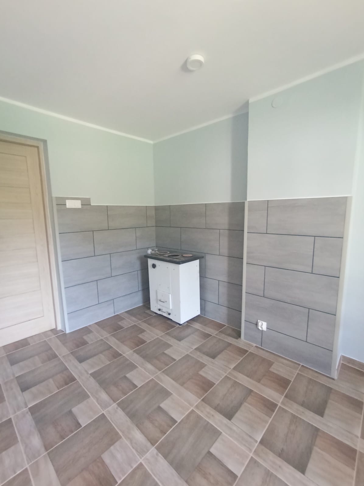 Wykończona kuchnia z szarymi płytkami imitującymi cegłę na ścianach do połowy wysokości, brązowymi płytkami podłogowymi ułożonymi w geometryczny wzór i białym piecem kaflowym narożnym.