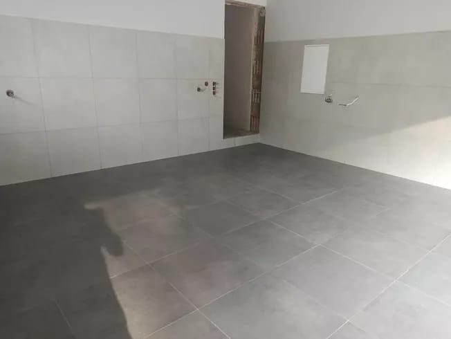 Pusta łazienka po remoncie z szarymi płytkami na podłodze i jasnymi płytkami na ścianach, widoczne wystające rury i otwór drzwiowy bez futryny.