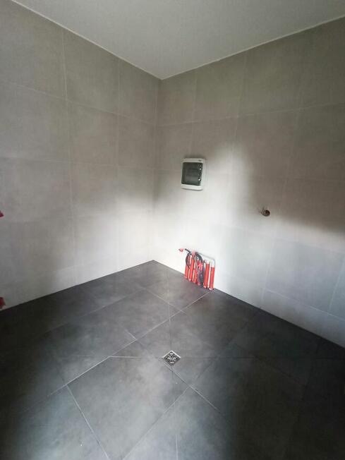 Wykafelkowana łazienka w trakcie remontu, widoczne szare płytki na ścianach i podłodze, rury instalacji wodnej w rogu, oraz kratka ściekowa w podłodze.