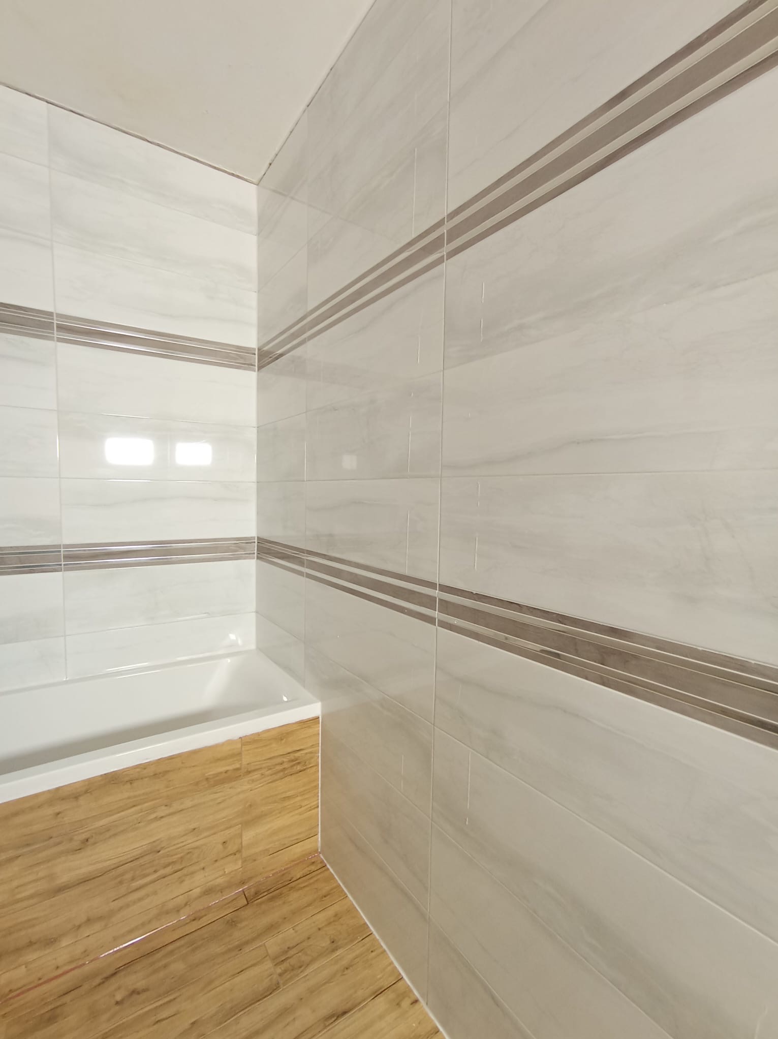 Wykończona łazienka z białymi płytkami imitującymi marmur, z poziomymi paskami w kolorze taupe, białą wanną i podłogą z paneli imitujących drewno.